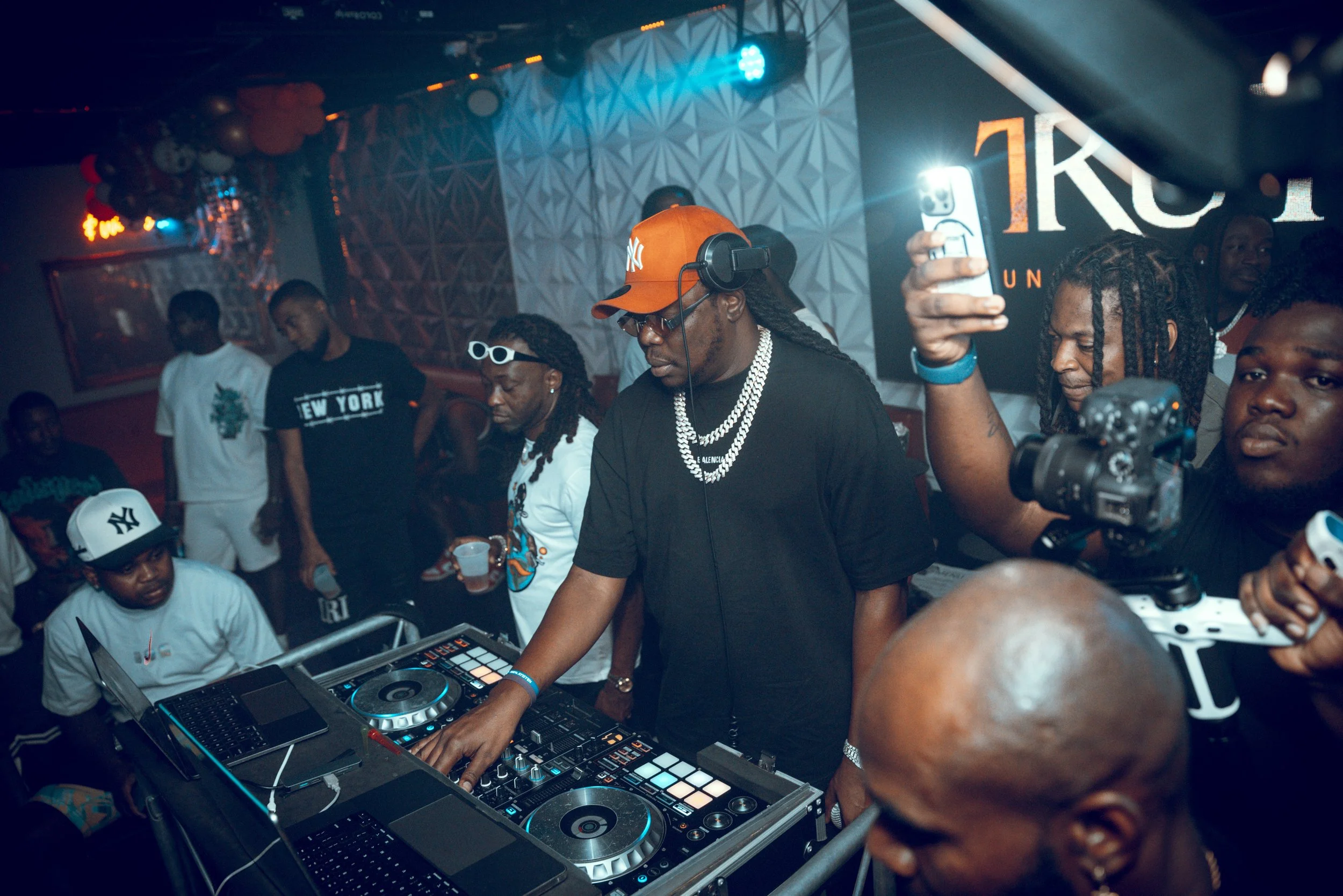 A DJ wearing a red cap and headphones is DJing at a club, surrounded by people, with some capturing the moment on their phones.