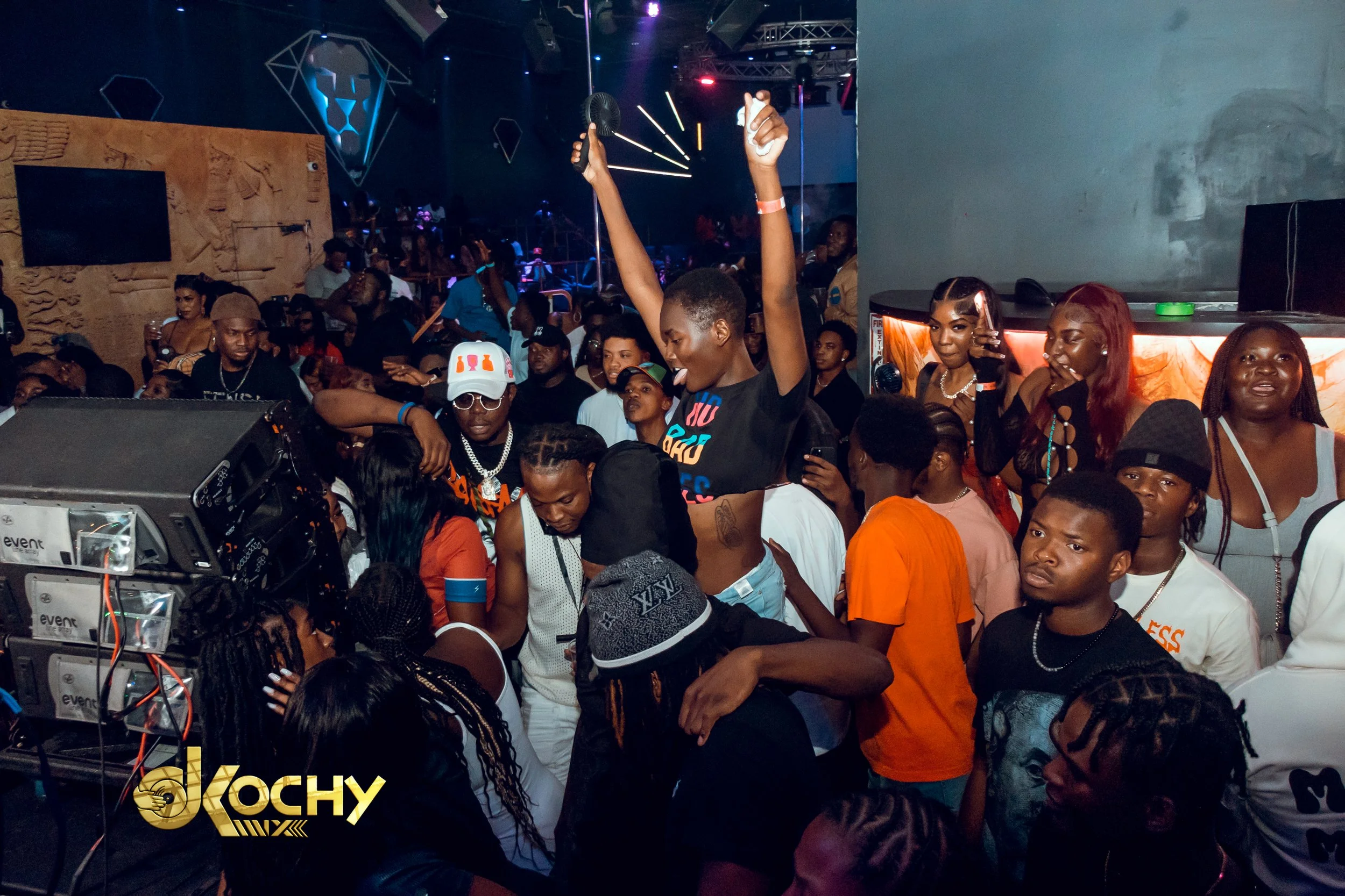 Crowd of people at a lively indoor event or nightclub, with one woman dancing on a platform, holding a fan in one hand, surrounded by others enjoying music and socializing.