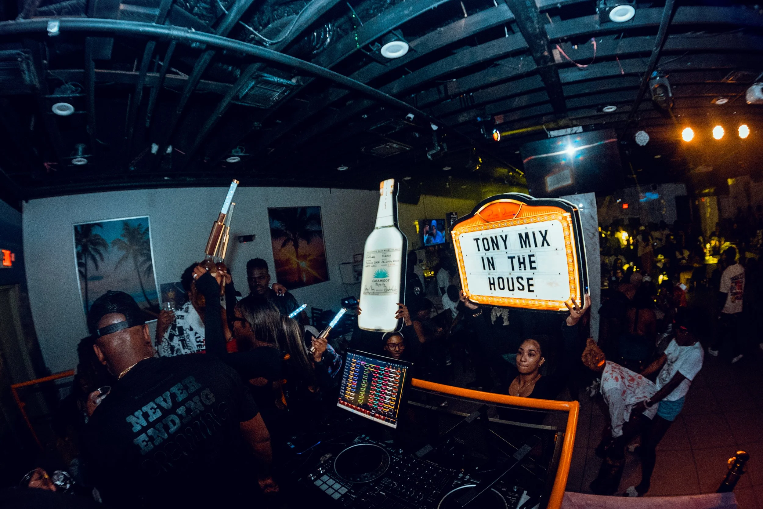Crowd enjoying a party at night with a DJ setup; large bottle cutout and sign reading 'Tony Mix in the House' are visible.