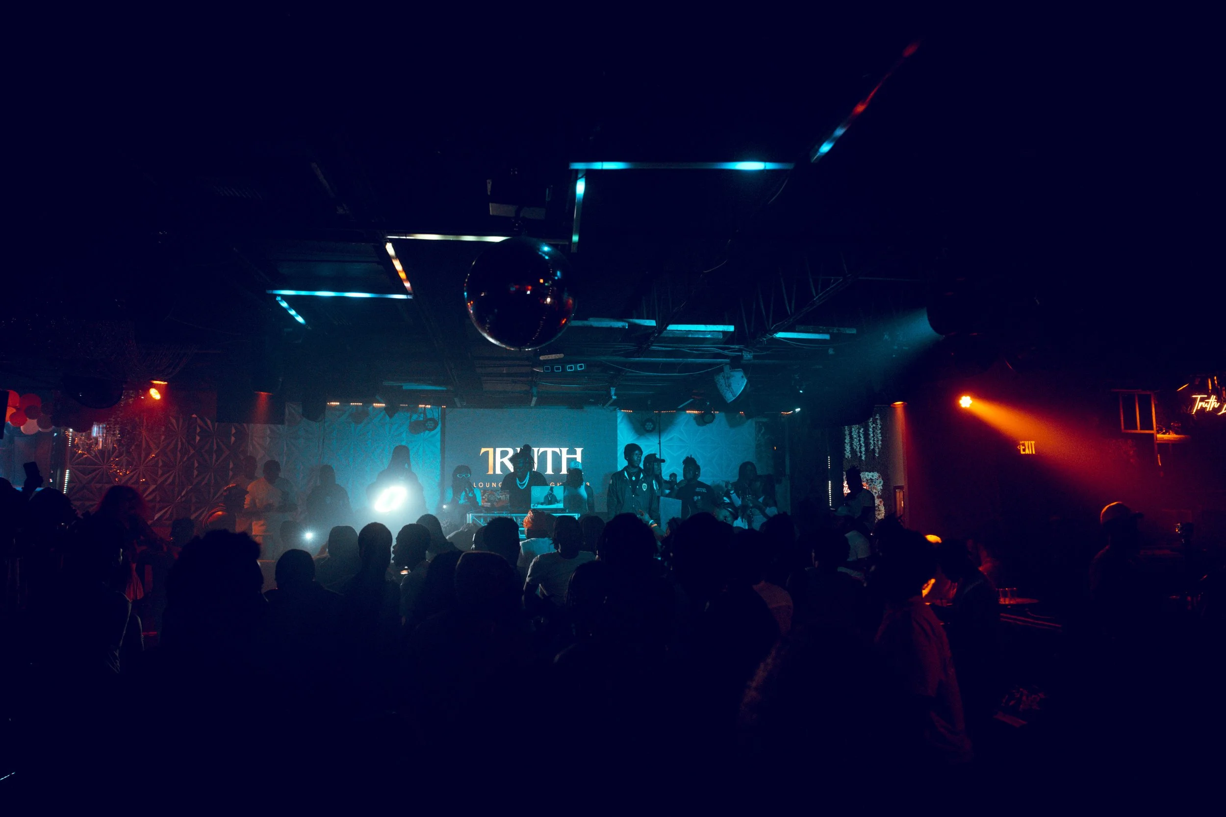 A nightclub scene with people dancing and socializing under colorful and dim lighting, with a stage featuring a band or performers, and a large screen displaying the word 'TRUTH'.