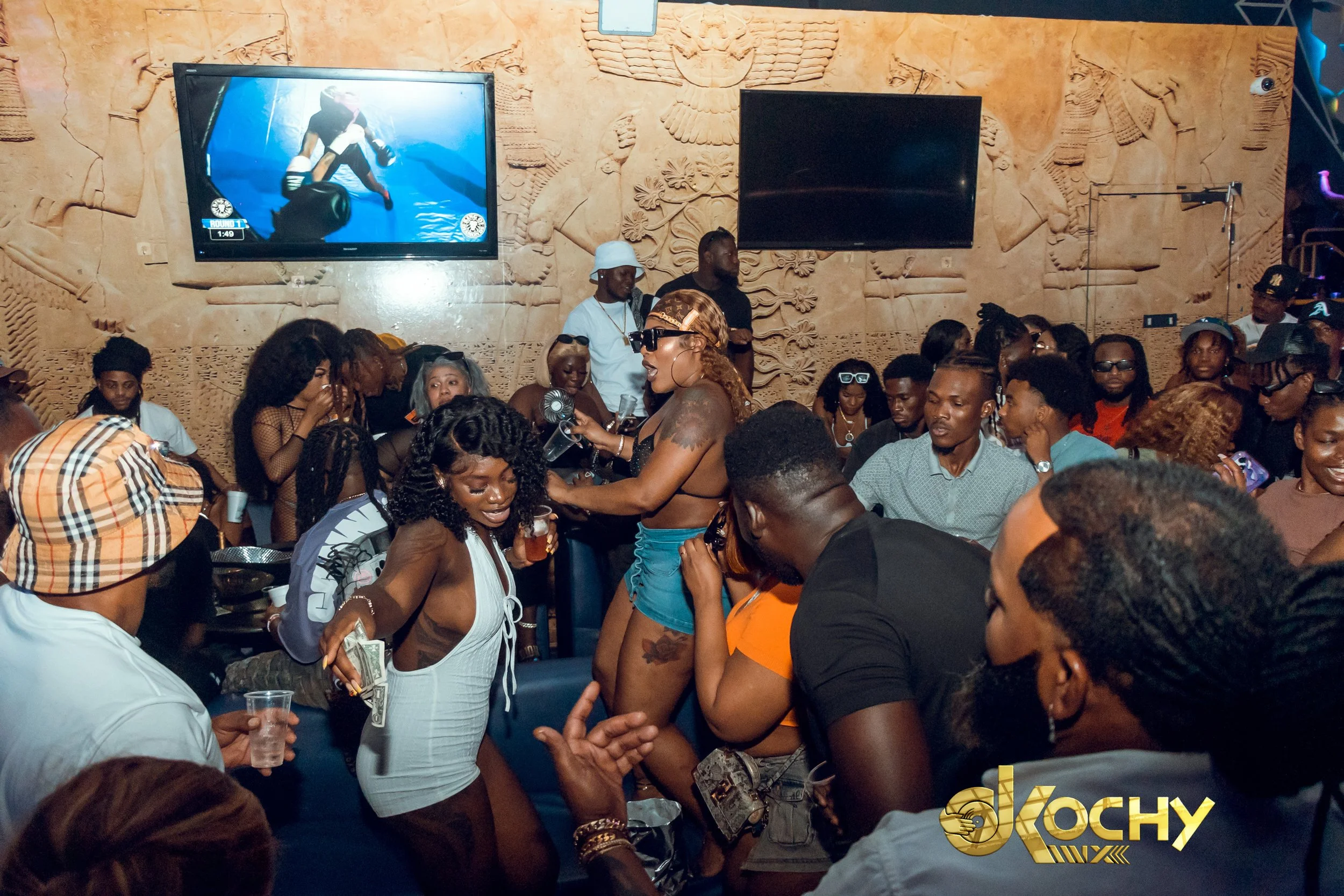 Crowd of people dancing and socializing in a lively indoor party with TV screens on the wall showing a boxing match, and an ornate carved wall behind them.