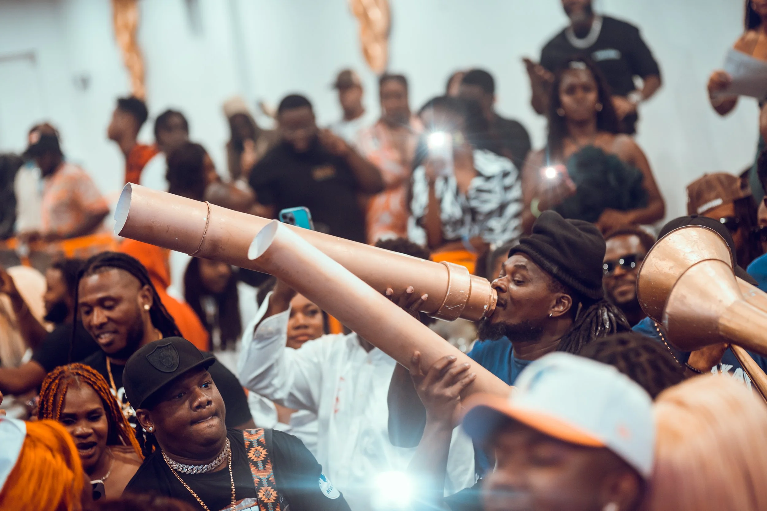 A man in a black hat and blue shirt is speaking into a large homemade telescope made from brown cardboard tubes at a crowded indoor event with many people watching.