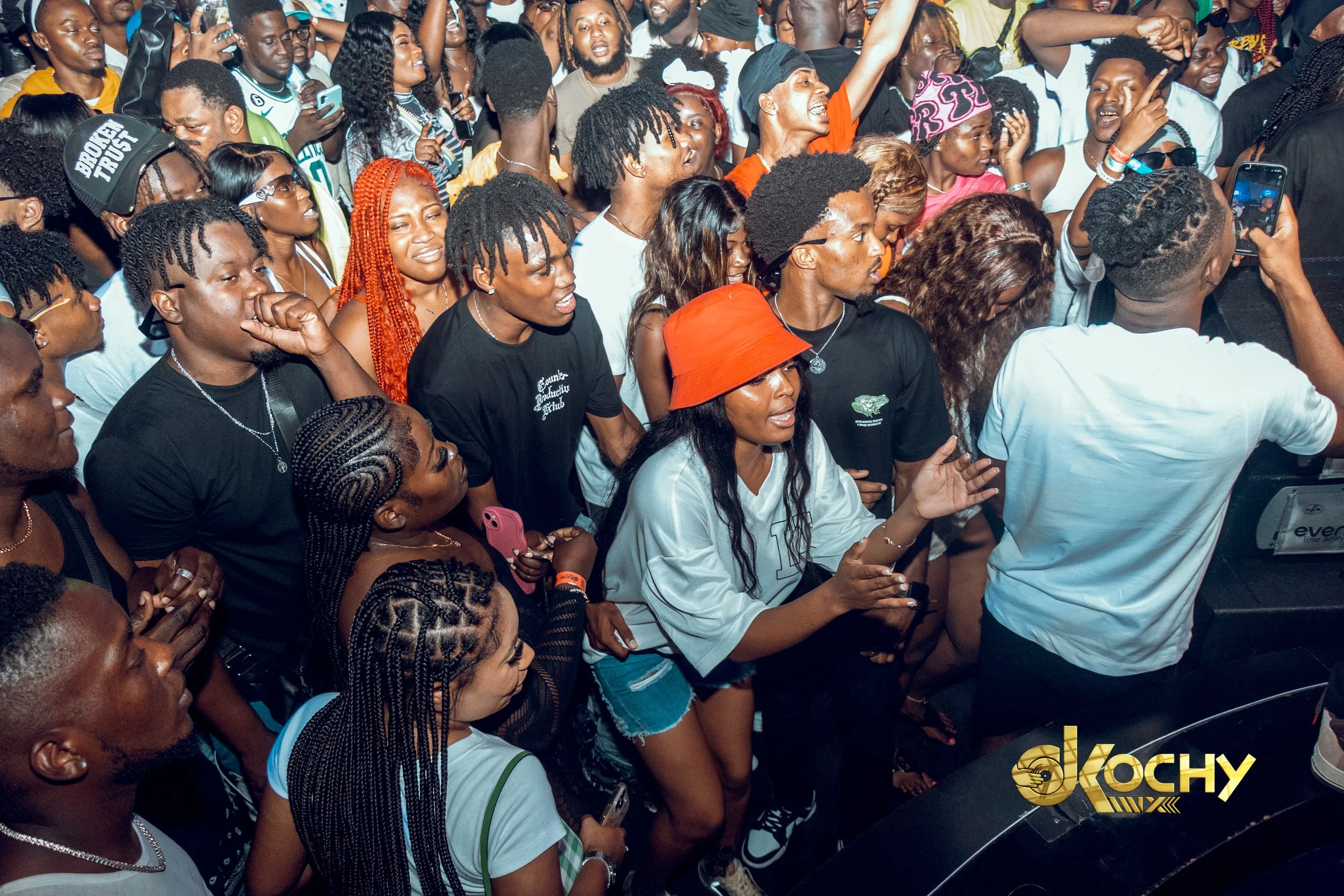 Crowd of people dancing and enjoying music at a party or concert, with some holding up phones. The scene is energetic and lively, with diverse individuals in casual clothing.