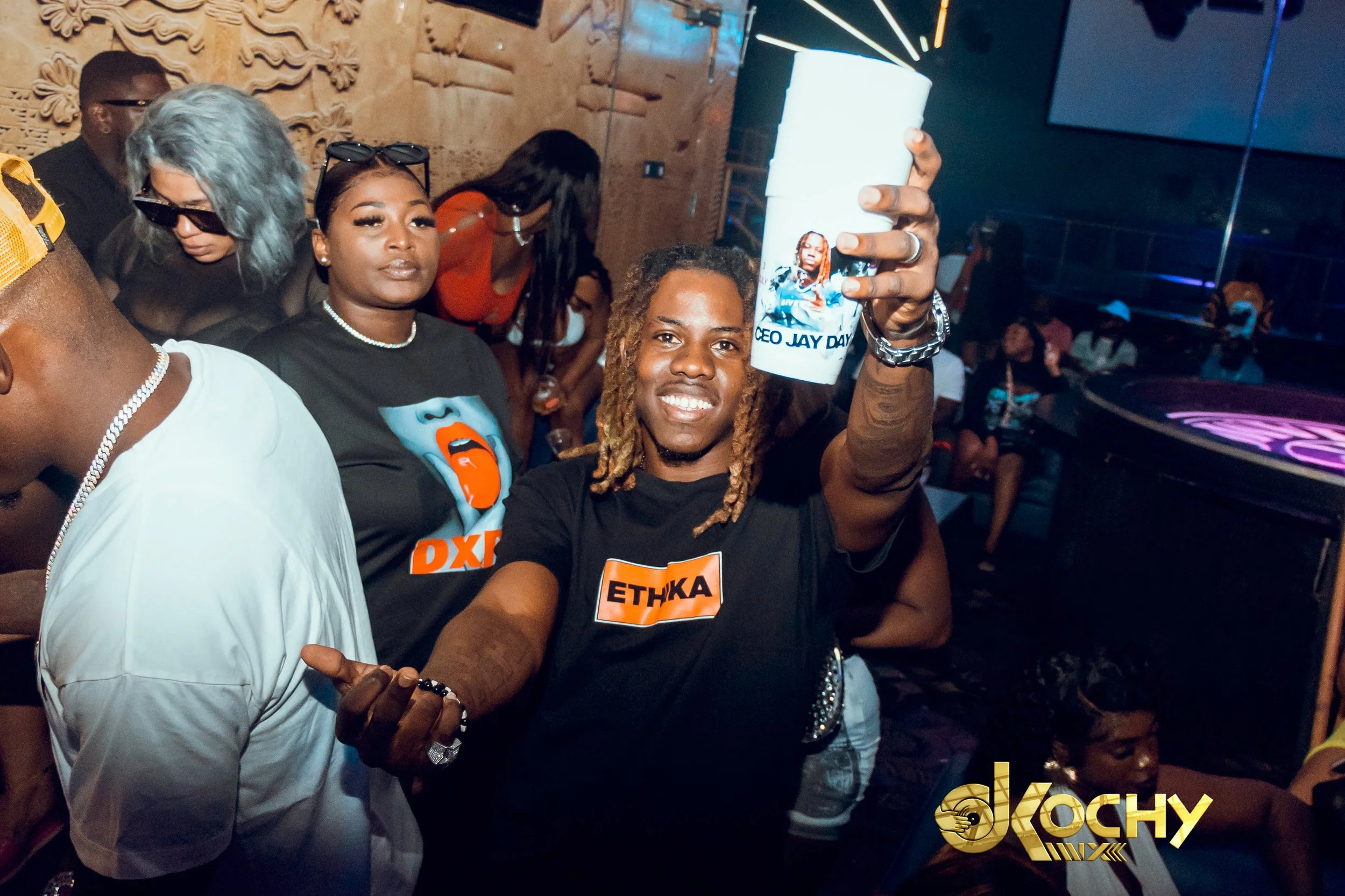 A man with dreadlocks smiling and holding a large white cup with a photo and text, at a lively party with other people dancing and socializing in a dimly lit venue.