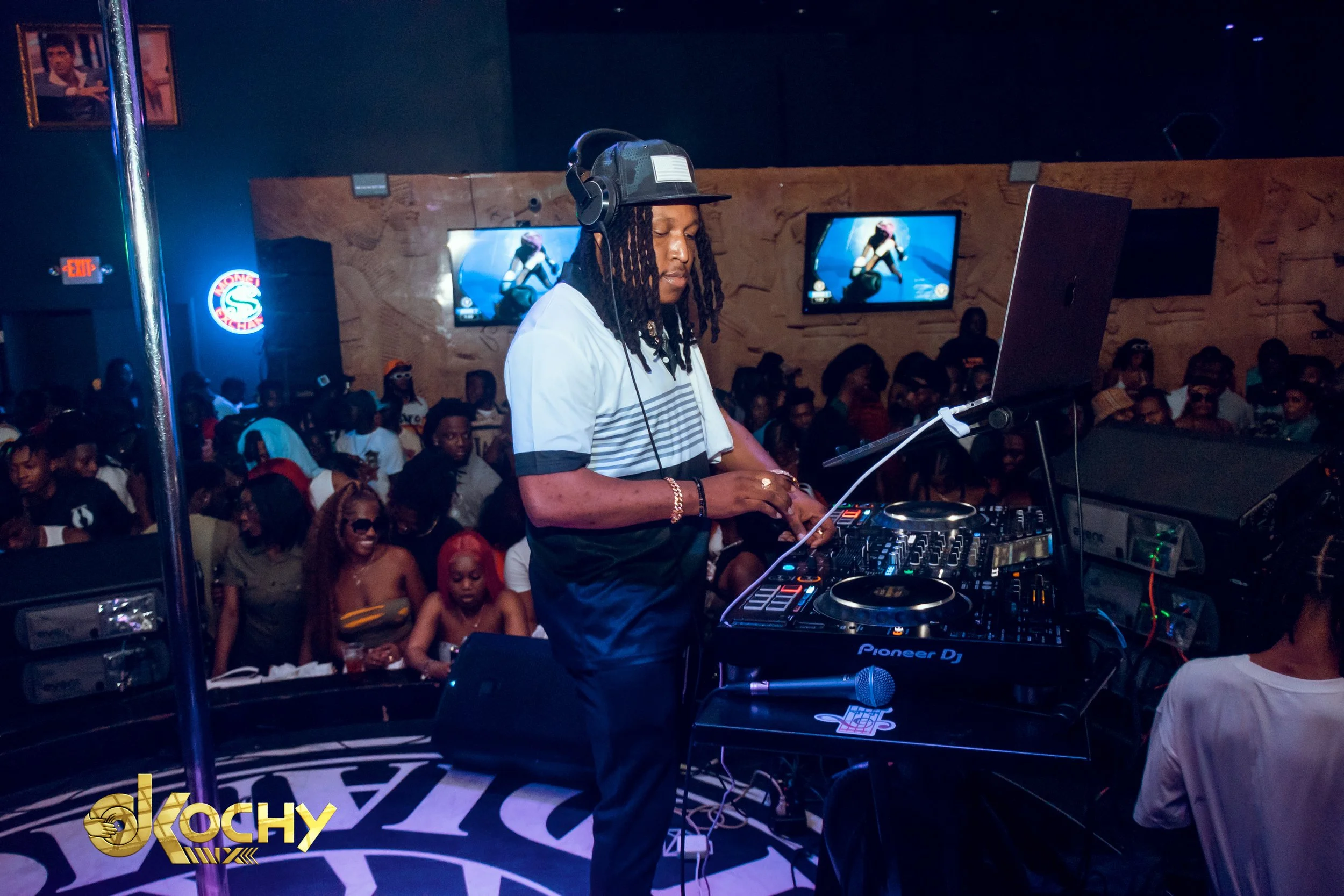 A DJ is performing at a nightclub, surrounded by a crowd of people. The DJ is wearing a striped shirt, a baseball cap, and headphones, standing at a DJ mixing console with a laptop, with screens in the background displaying an animated image.