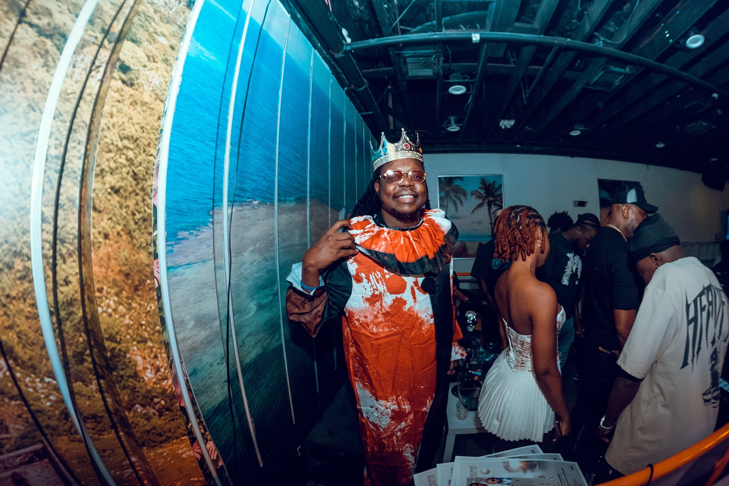 Man wearing a crown and a colorful outfit, smiling at the camera, at a party with other people, with a tropical ocean scene decor on the wall behind him.
