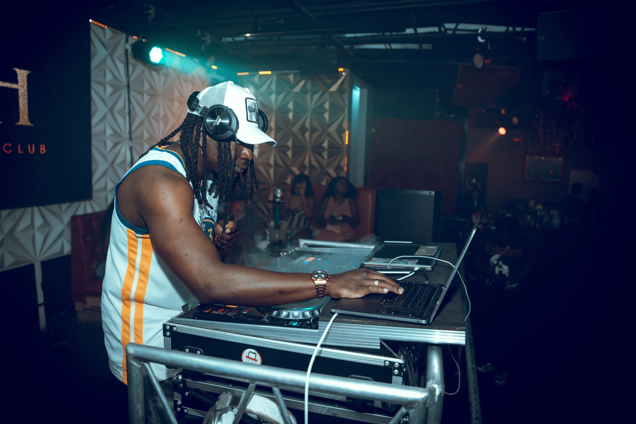 A DJ wearing a white baseball cap, headphones, and a sleeveless sports jersey is performing at a nightclub, standing behind DJ equipment and a laptop, with a dimly lit background and some people sitting in the distance.