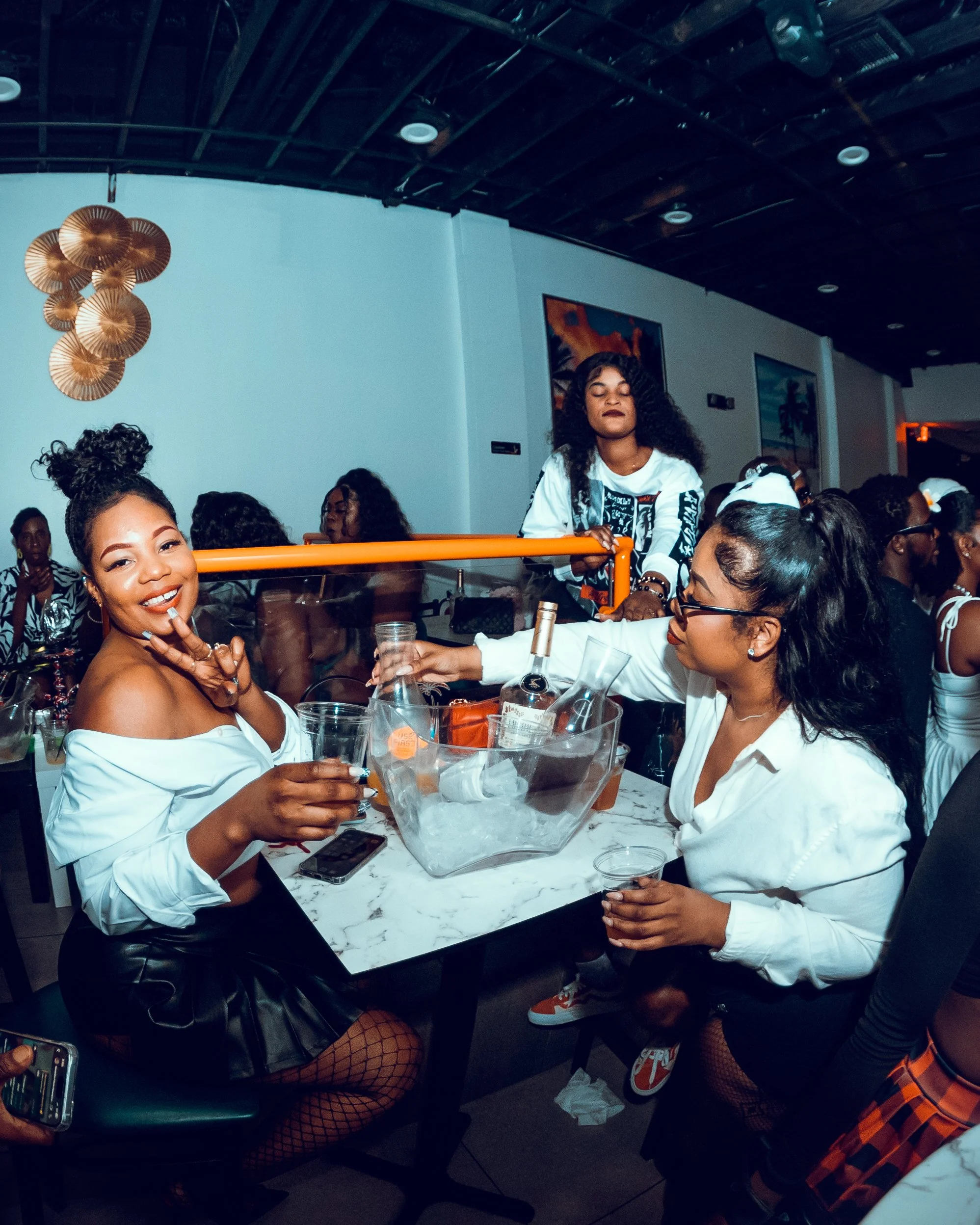 A group of women enjoying drinks at a party, with one woman smiling and making a peace sign at the camera while others are conversing and serving drinks.
