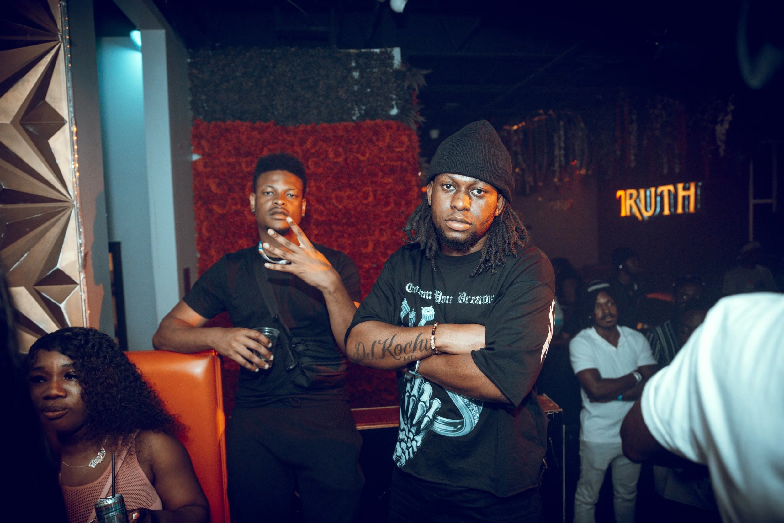 Two young men standing in a dimly lit nightclub, one wearing a beanie and with tattoos on his arm, the other with short curly hair and holding a drink, with several people in the background and a illuminated sign reading 'TRUTH'.