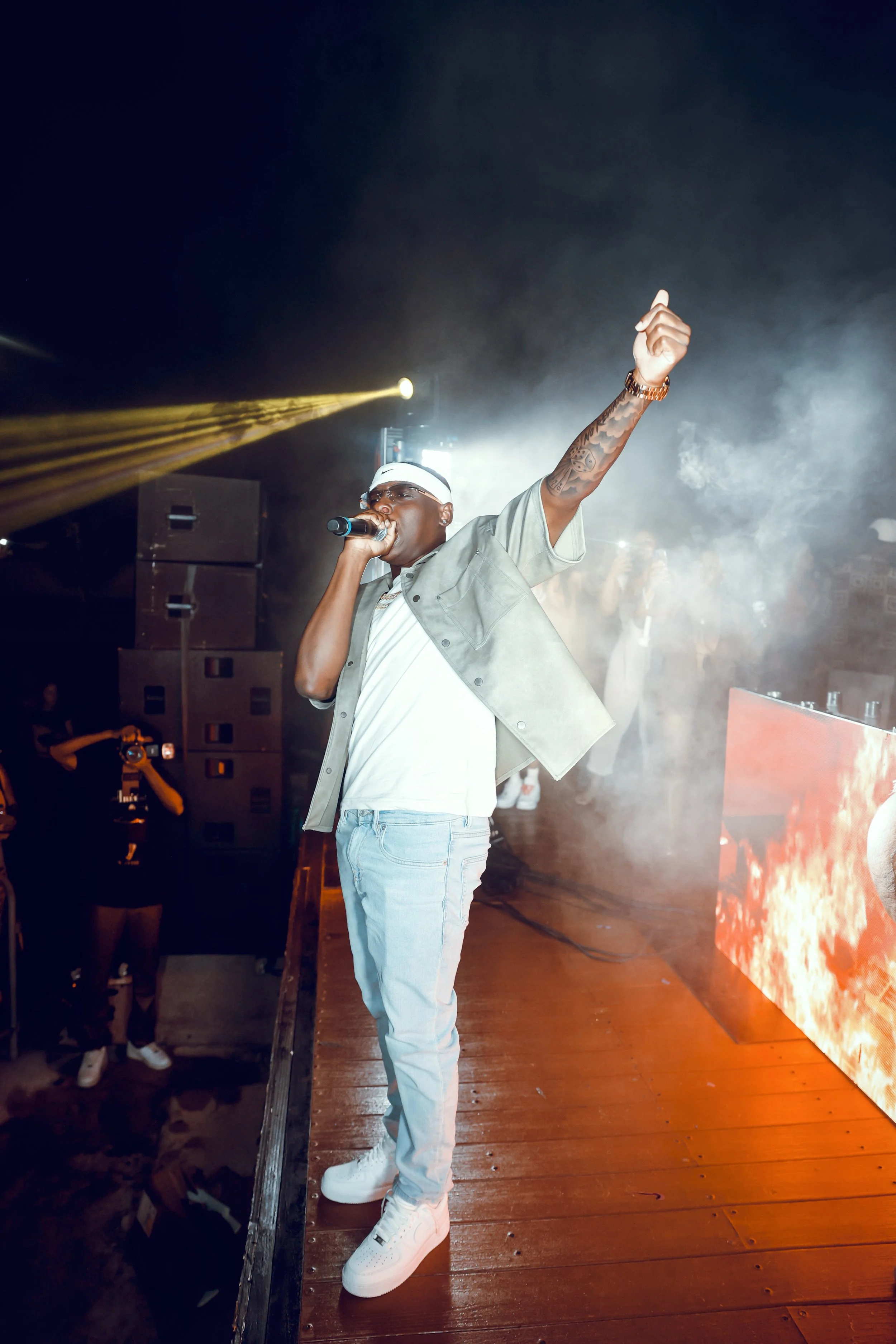 A male performer on stage holding a microphone and raising his hand, with bright stage lights and a smoky atmosphere in the background.