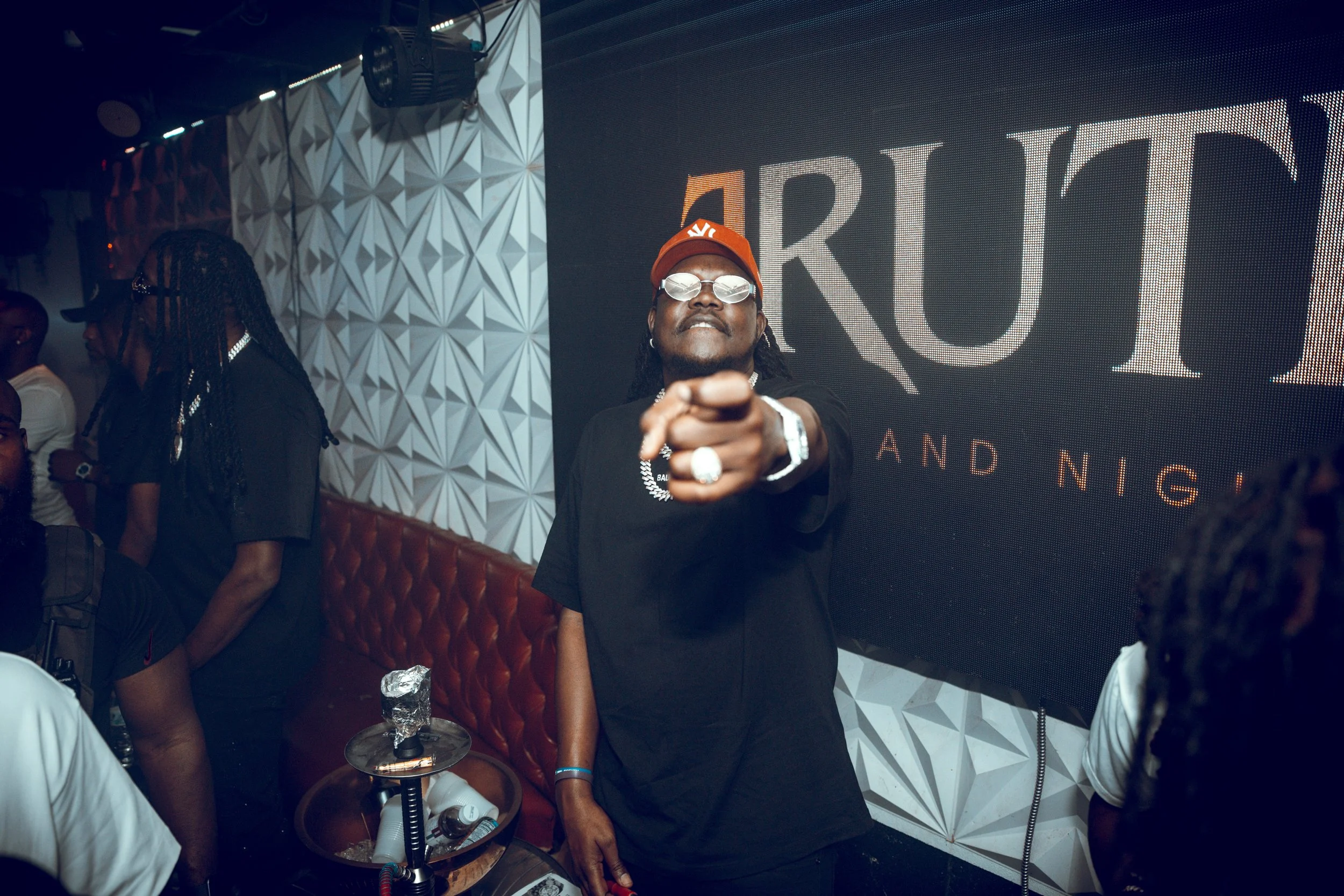 Person wearing sunglasses, a red New York Yankees cap, and jewelry, pointing towards the camera in a nightclub with textured wall and large digital screen in the background.