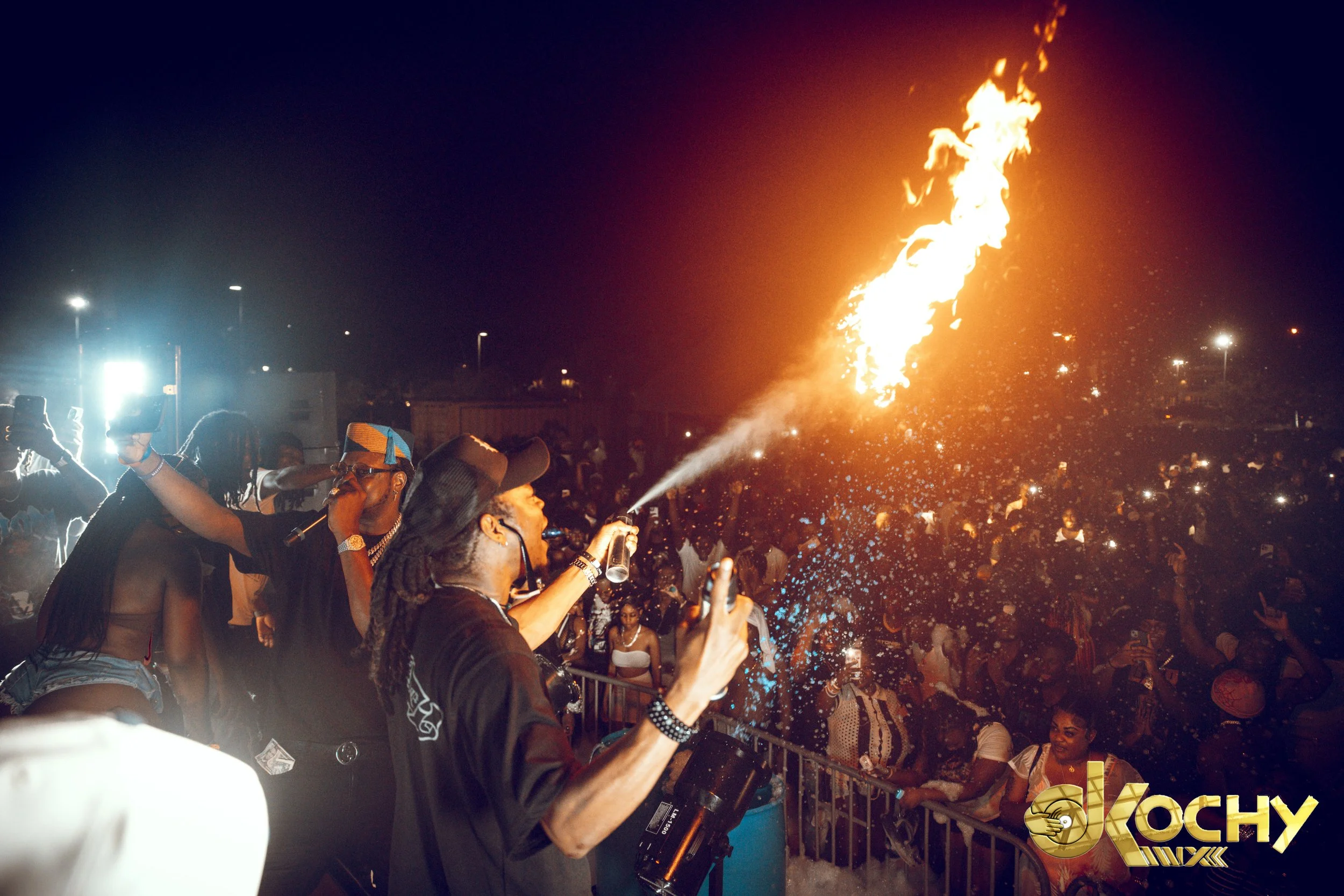 A DJ and a person with dreadlocks and glasses performing in front of a crowd at a night party, with flames and sparks in the background.