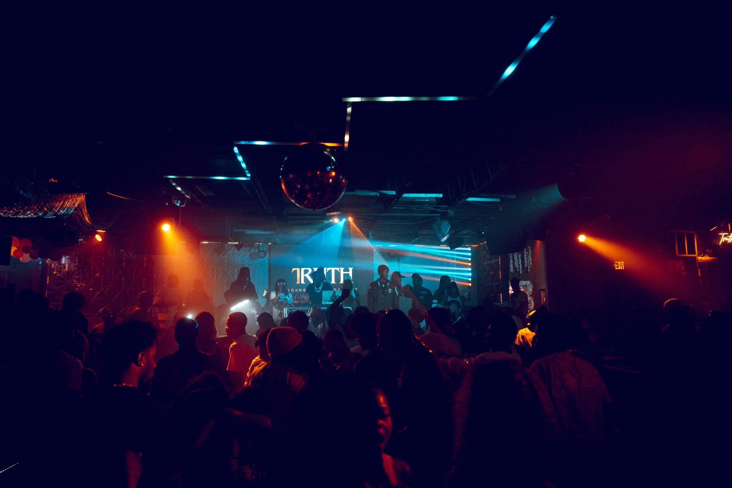 Nightclub scene with a stage, colorful lights, a DJ, and a crowd dancing.