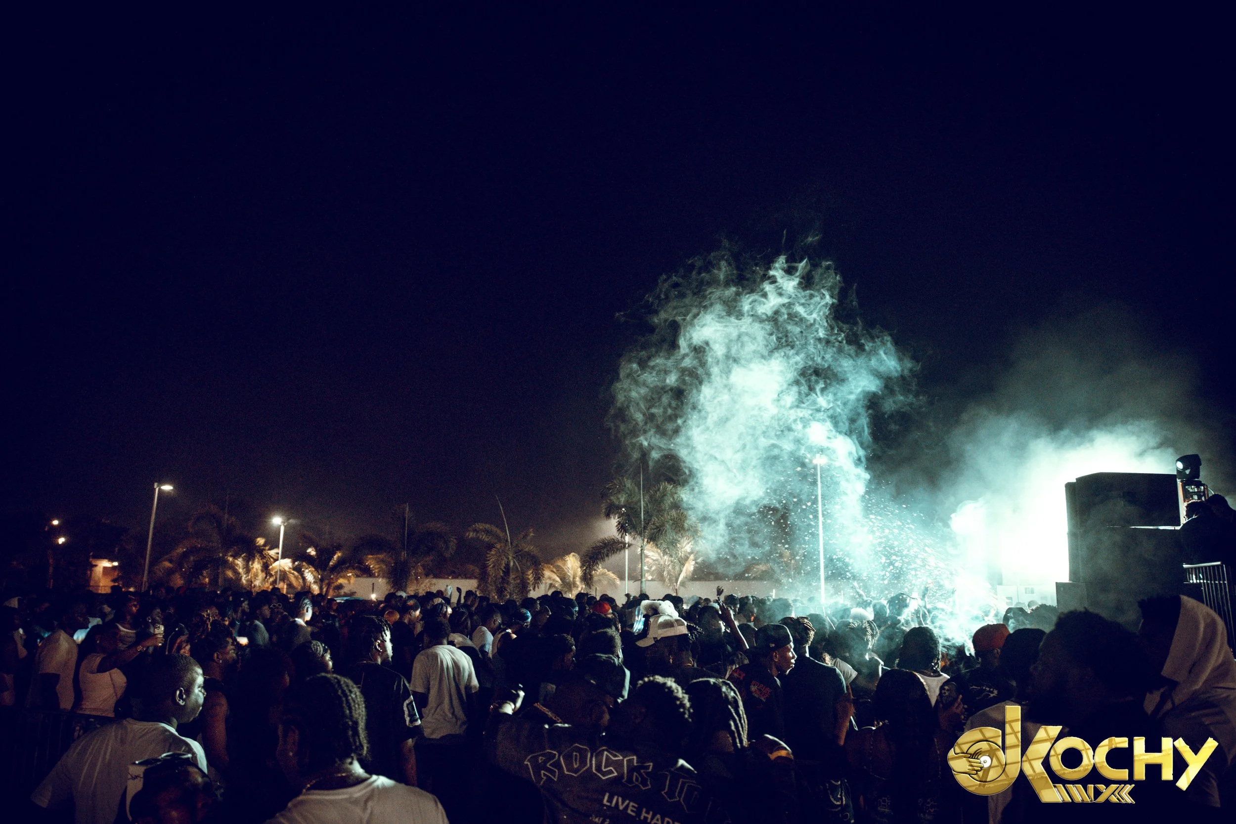 Crowd of people at night watching a fireworks display or light show, with smoke and bright light in the sky, palm trees in the background, and some people taking photos.