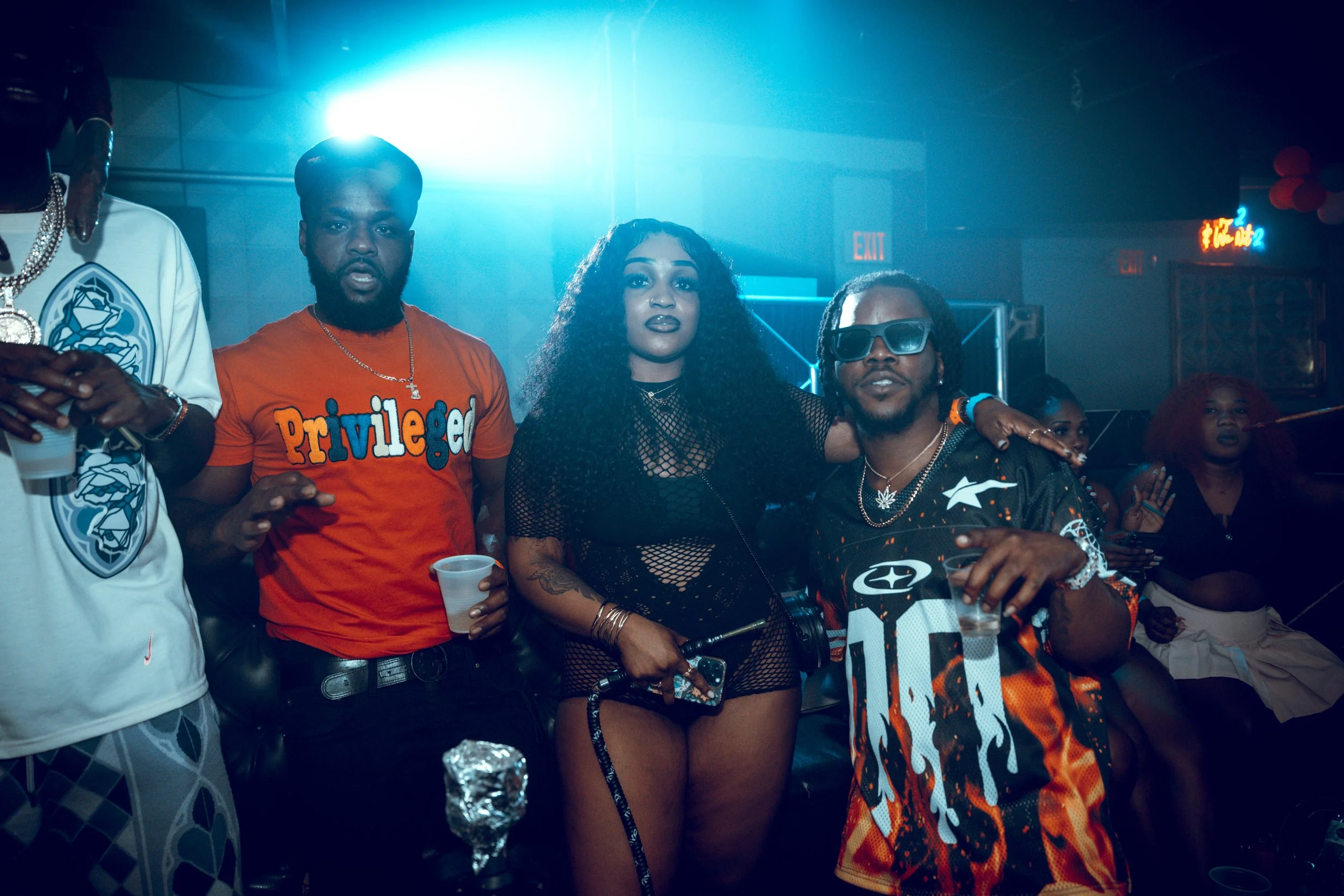 Group of people at a party, including a woman with curly hair in black fishnet clothing and sunglasses, and men wearing colorful shirts, holding drinks, in a dimly lit club with vibrant blue lighting.