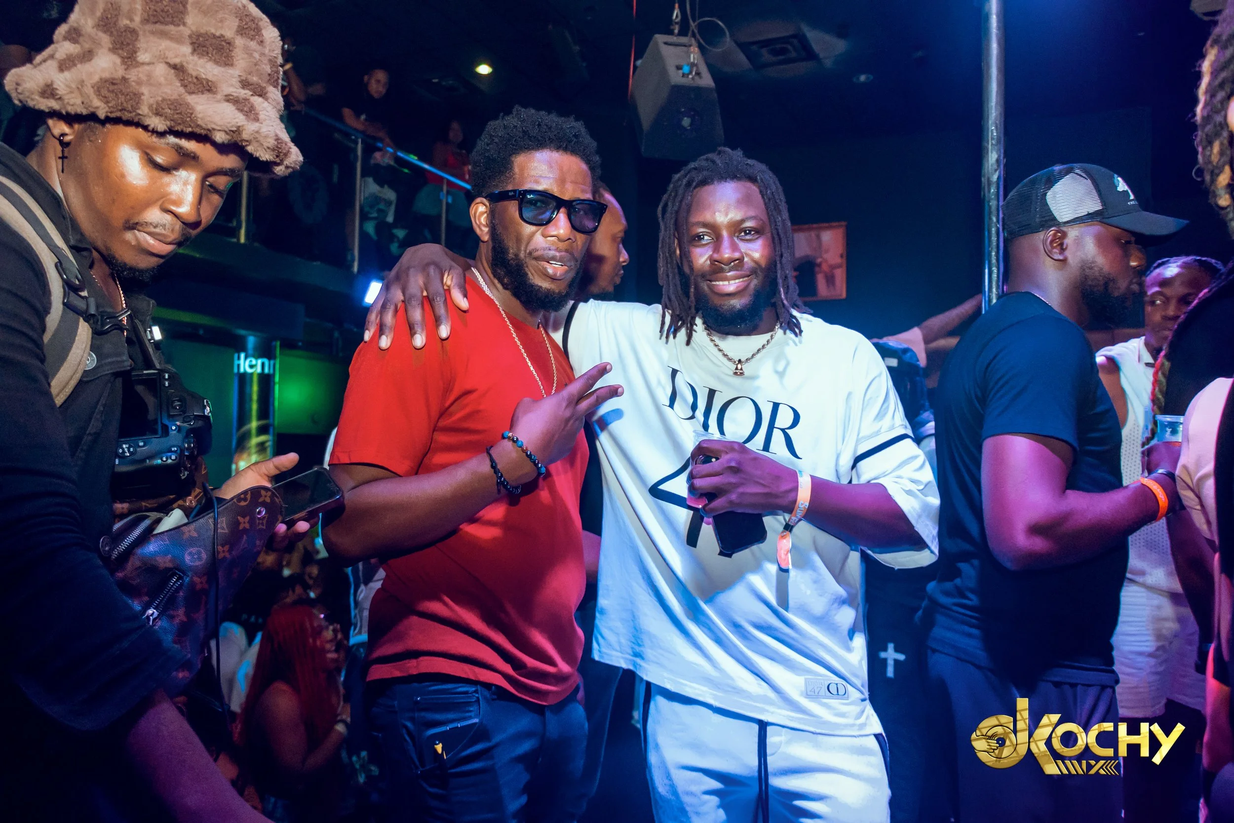 Two men are posing together at a nightclub, one wearing a red T-shirt and sunglasses, and the other in a white Dior T-shirt. They are smiling and making hand gestures, surrounded by other people in a lively setting with colorful lighting.