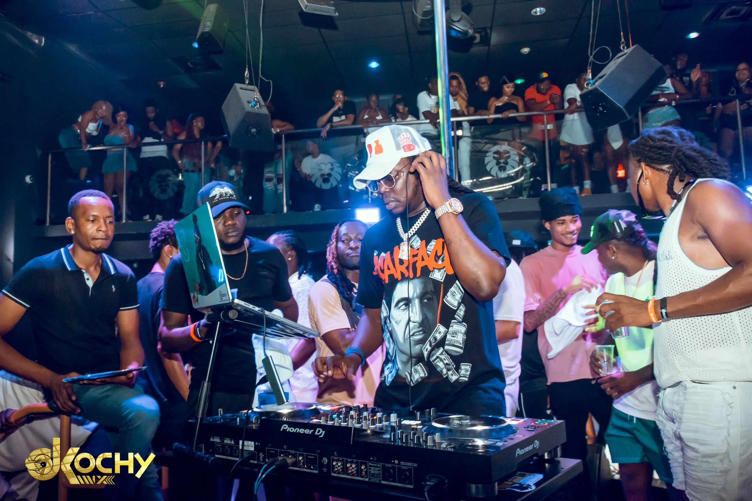 A DJ wearing a black shirt with a graphic of a woman and the word 'SHARFAY', a white cap, glasses, and jewelry, is performing at a nightclub surrounded by a diverse group of people dancing and socializing. The club has dim lighting with multiple peop