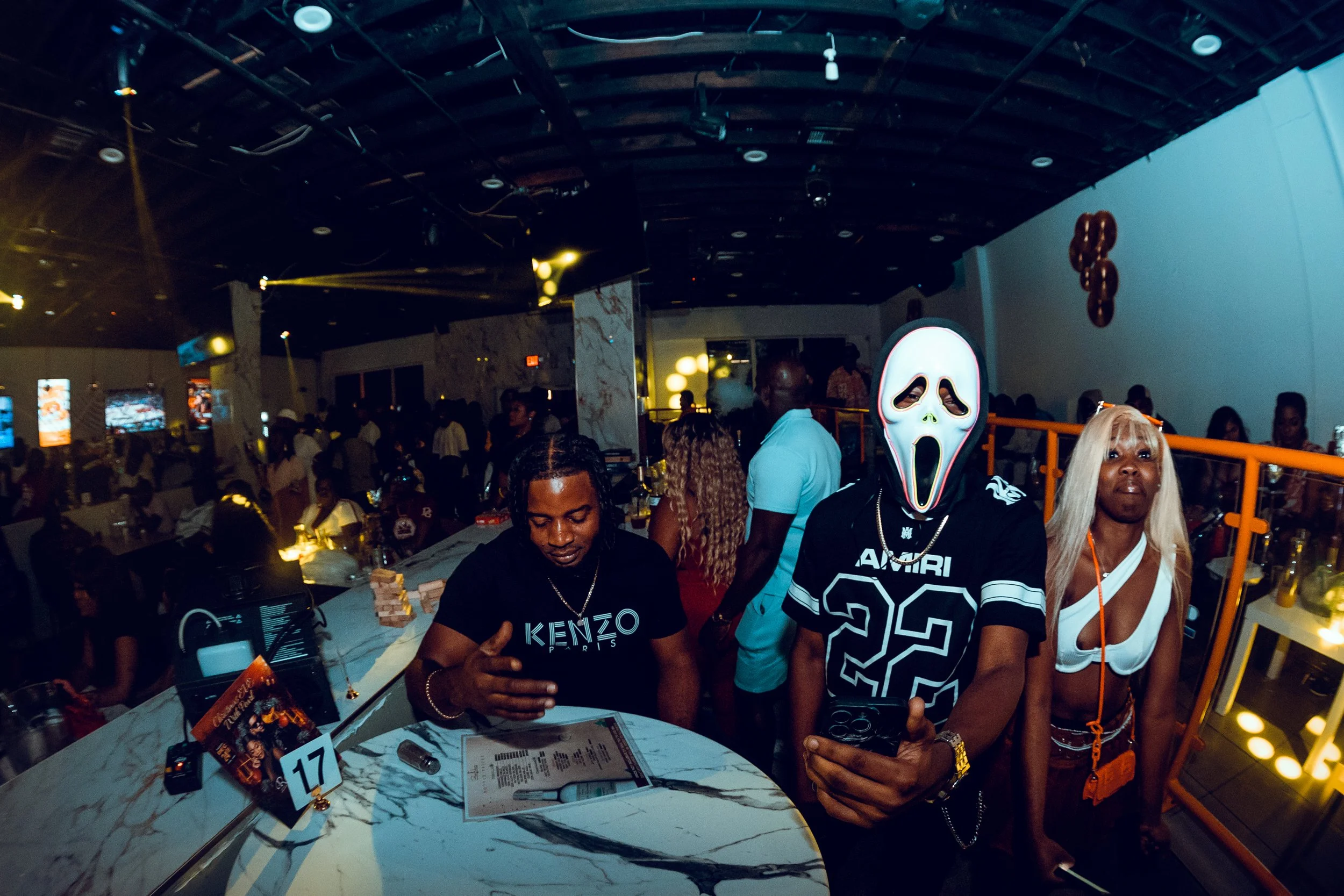 People at a party or nightclub, one person wearing a ghost face mask from the movie Scream, others are dressed casually, some looking at their phones, with a lively crowd in the background.