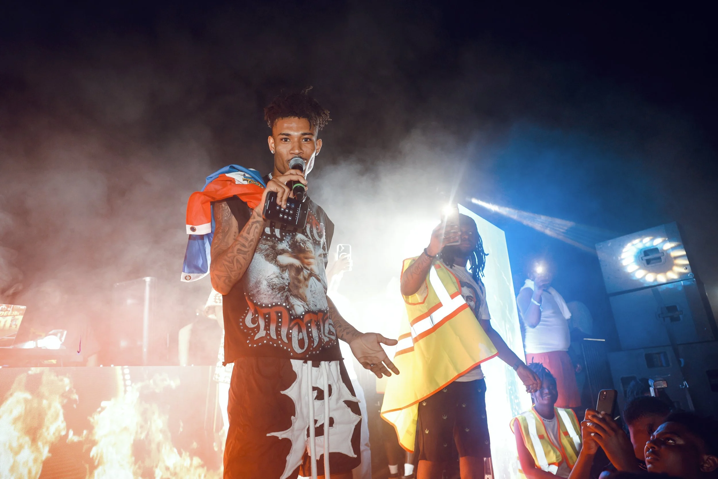 A performer with a microphone on stage, wearing a graphic t-shirt, with an Israel flag draped over shoulders, and others in safety vests in the background, stage lights and smoke.