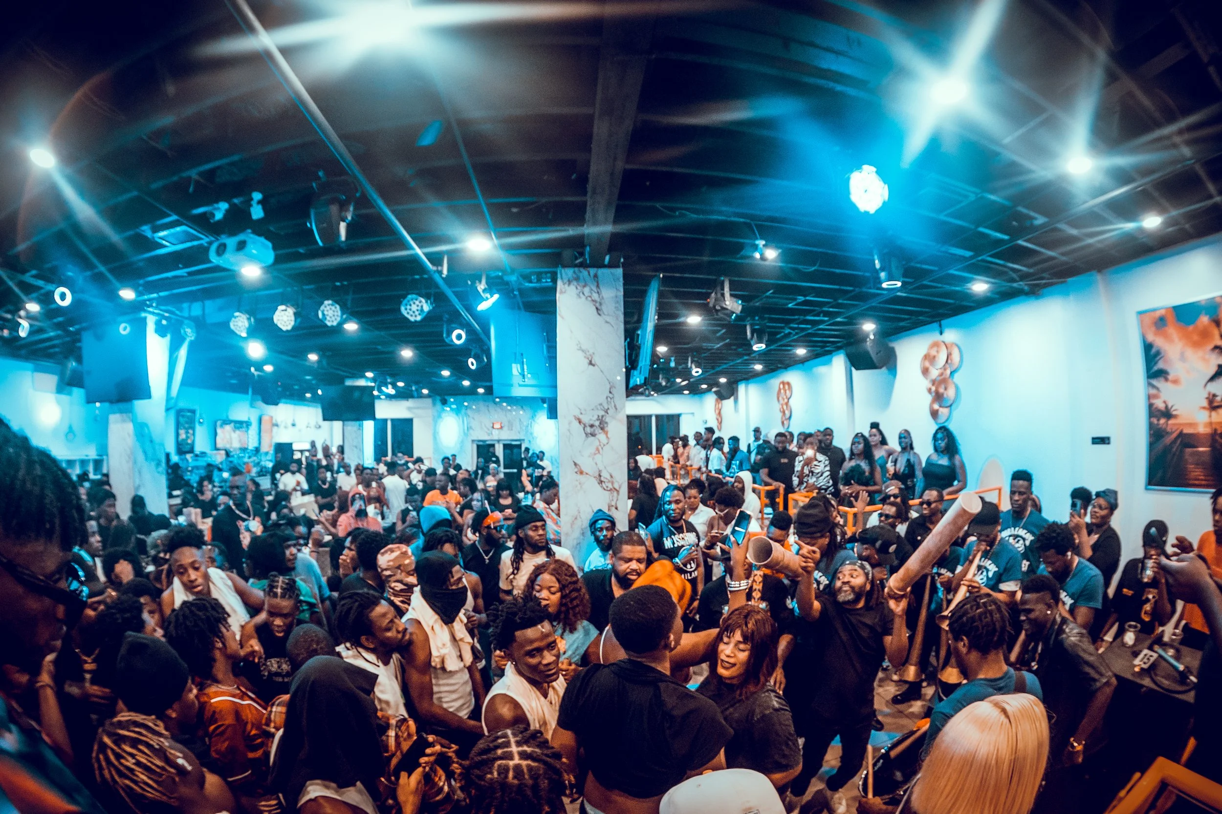 Large indoor dance party with a crowd of people dancing and socializing under vibrant blue and orange lighting, with music equipment and artwork on the walls.