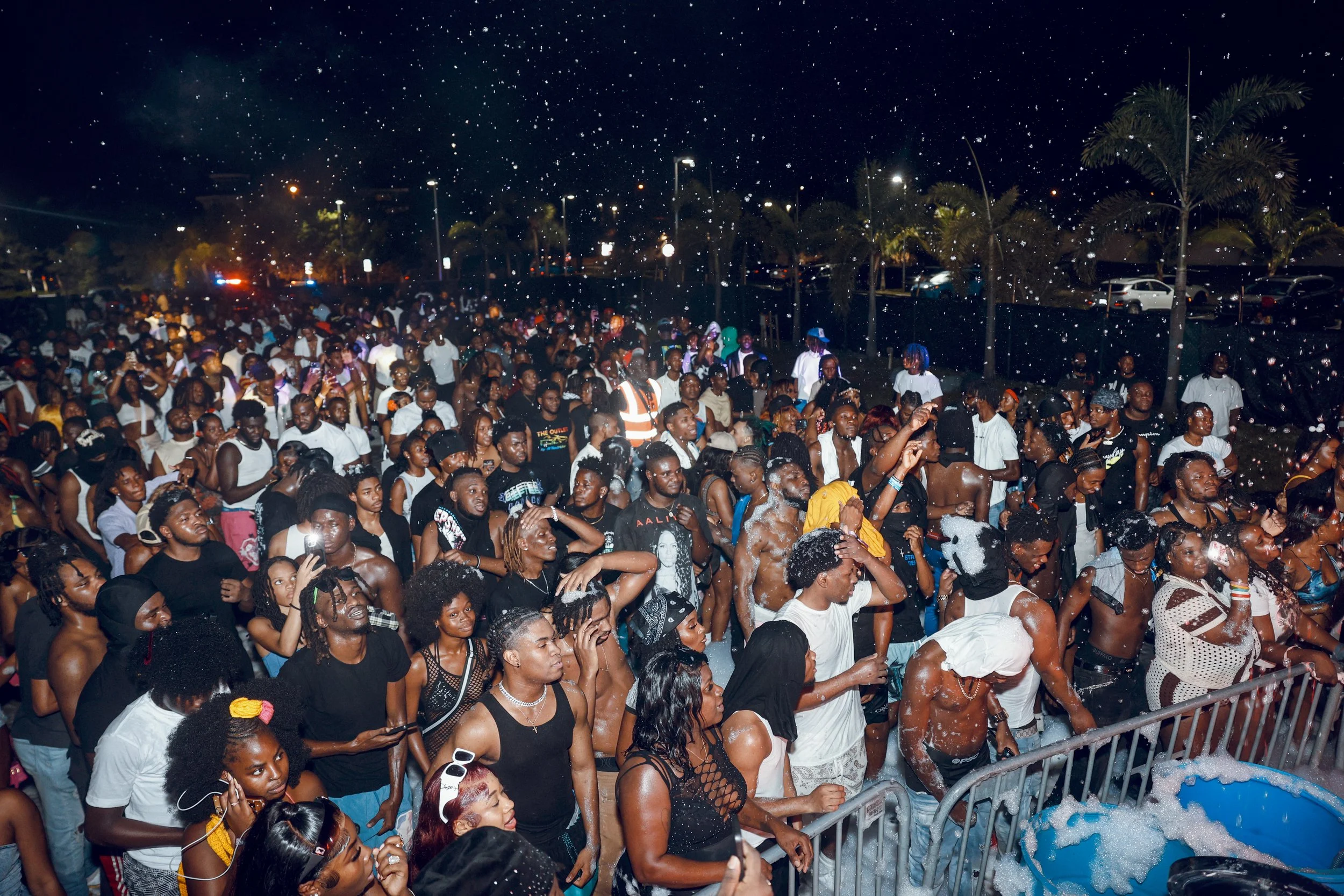 Crowd of people at a nighttime outdoor event, some are taking photos, others are dancing or watching, with foam on the ground and palm trees in the background.