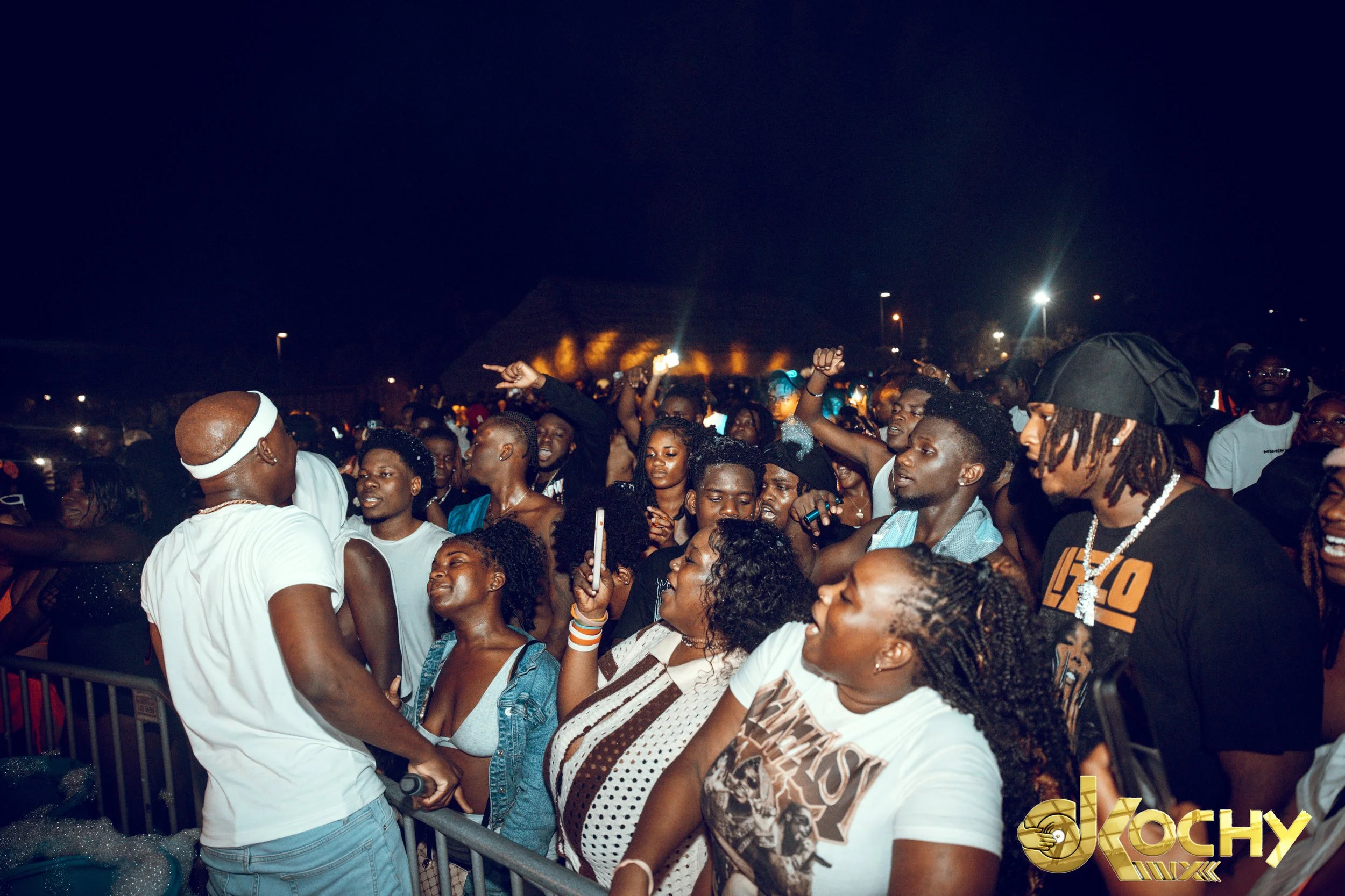 Crowd of people at an outdoor concert or event at night, some taking photos, dancing, and enjoying the music, with stage lights in the background.
