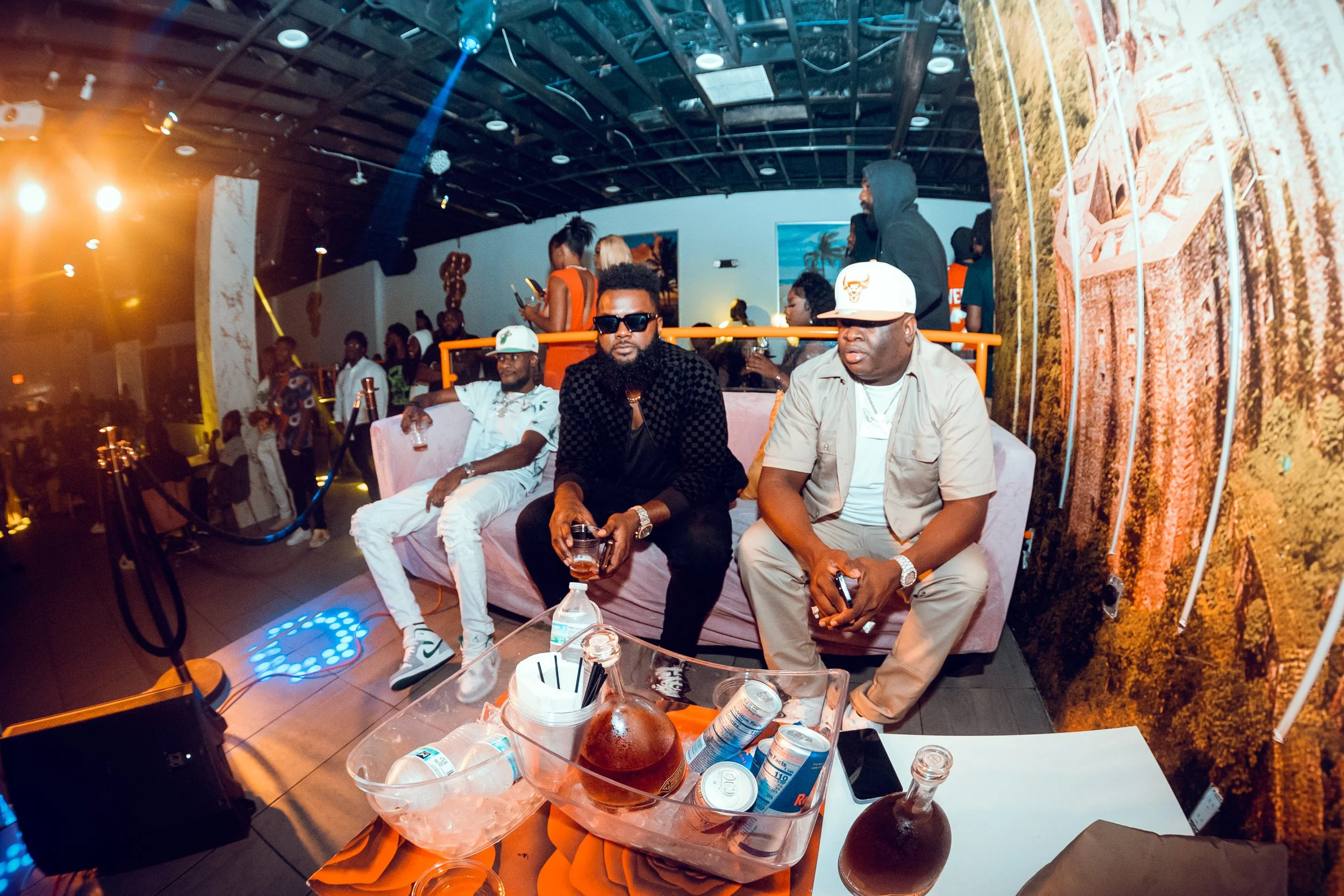 Three men sitting on a pink couch at a party or event. The man on the left is dressed in white, the man in the middle is wearing black with sunglasses, and the man on the right is in tan with a white hat. There are drinks on a table in front of them,