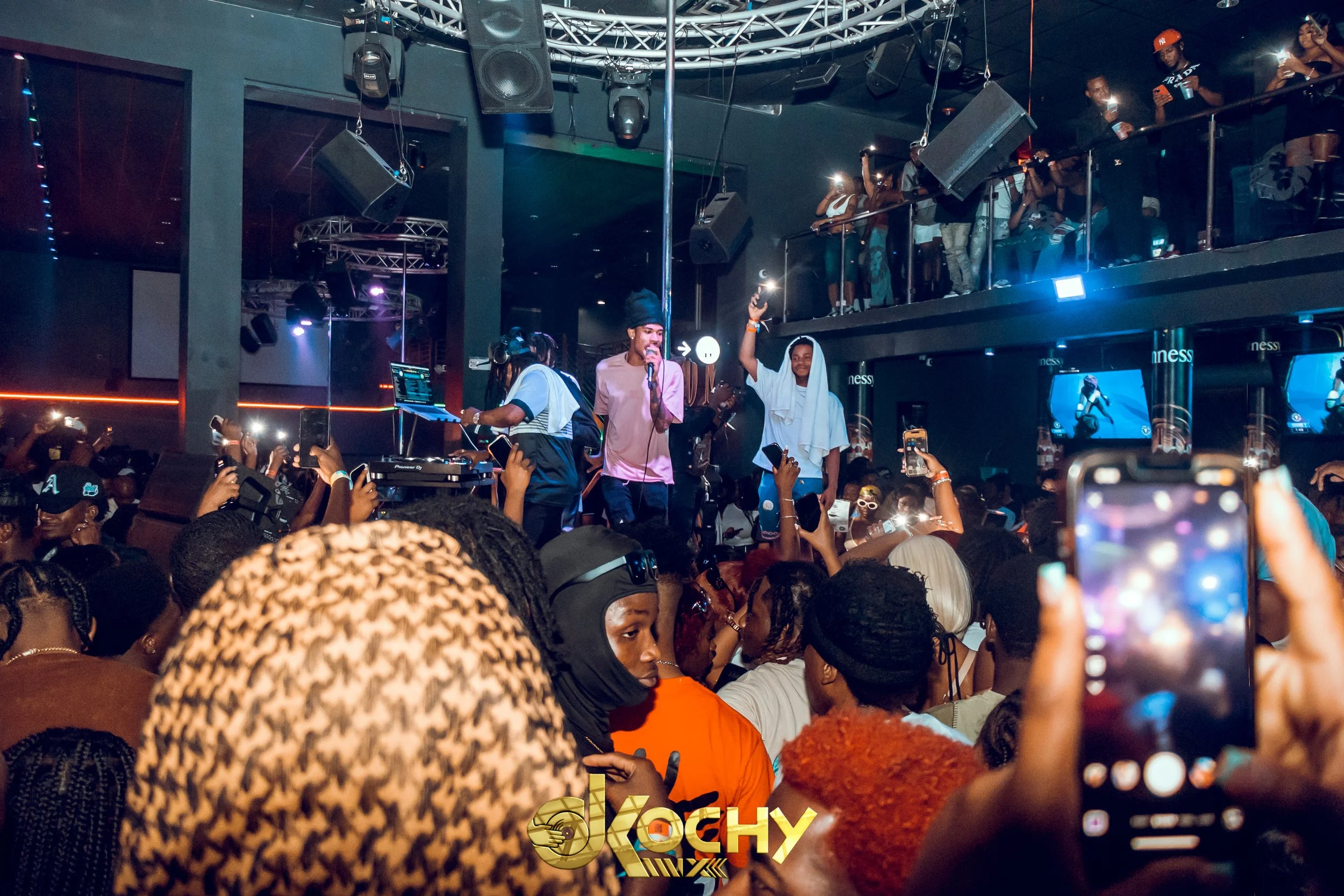 A crowded nightclub with a live performance on stage. A male singer in pink shirt holding a microphone, and another man in white with a towel on his shoulders holding a microphone. A DJ is on the left side of the stage, surrounded by people taking ph