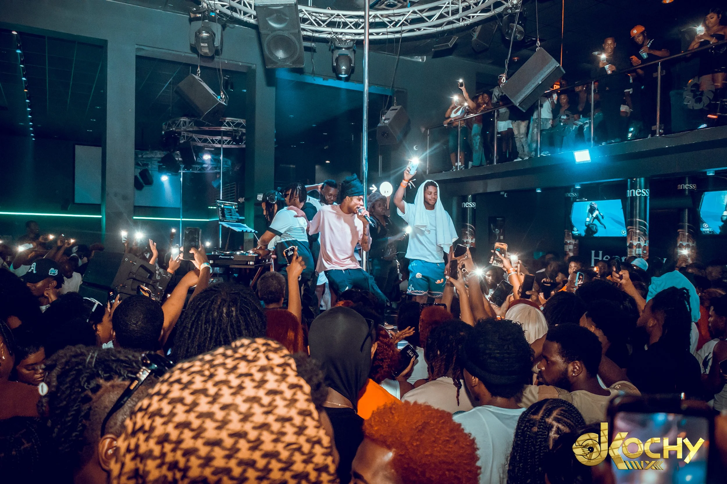 A lively indoor concert scene with performers on stage and a large crowd of fans taking photos, many holding up their phones with briefs flashes of light. There is a DJ setup in front of the stage, and some audience members are on an upper balcony wa