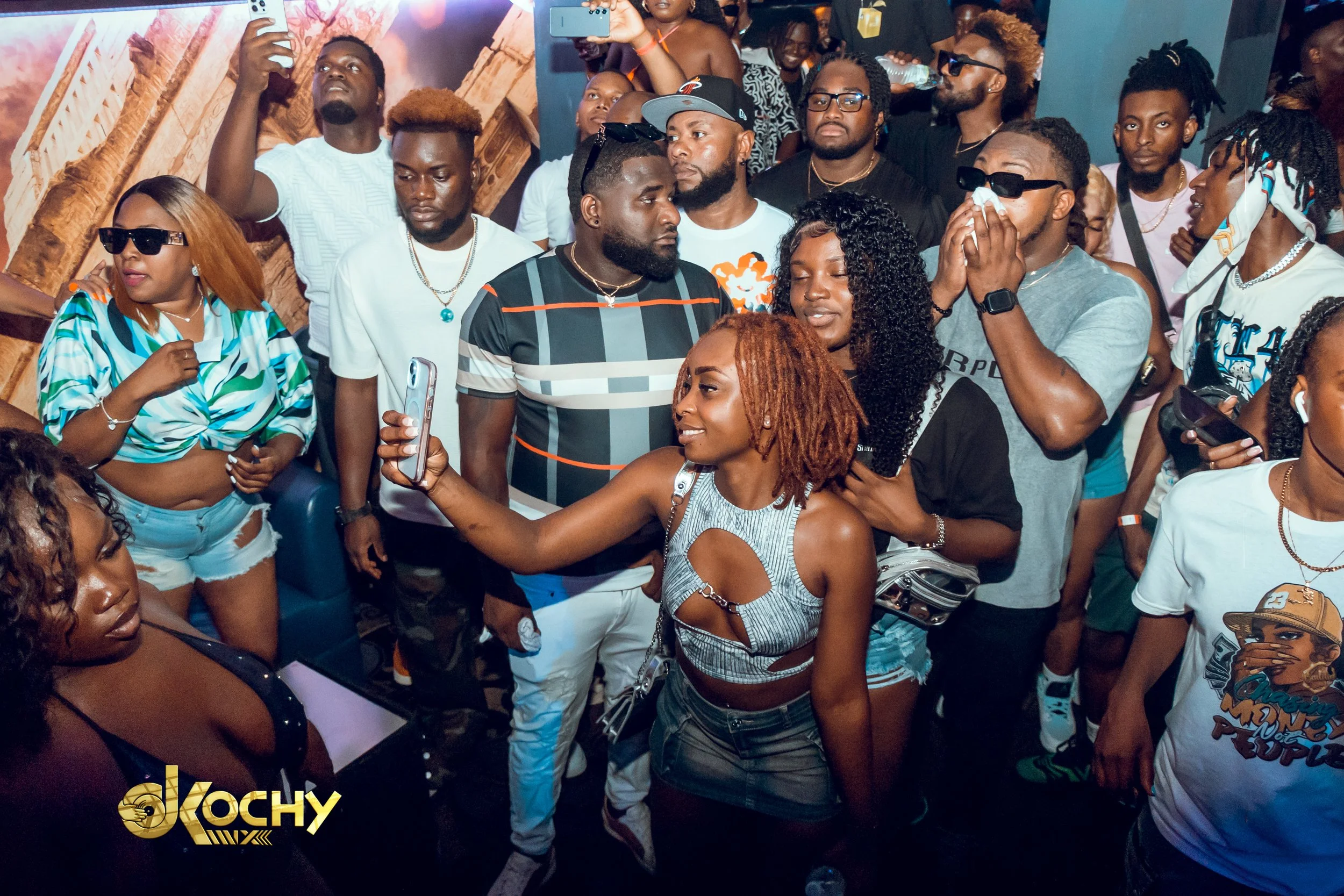 Group of people at a lively event, taking selfies and watching entertainment with some wearing sunglasses and casual clothing.