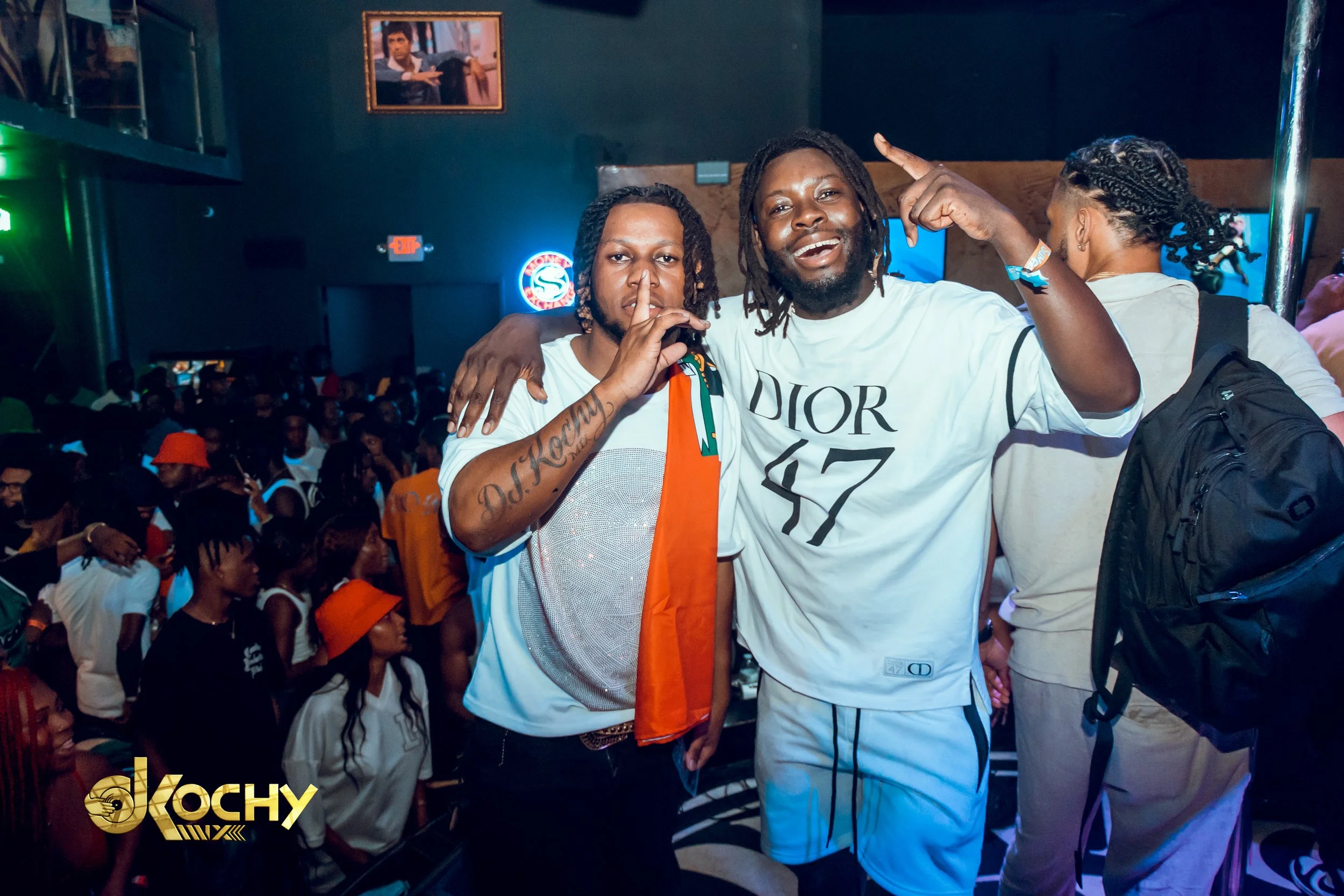 Two men celebrating at a crowded party, one making a shushing gesture and the other smiling and pointing up, with a lively crowd and neon signs in the background.