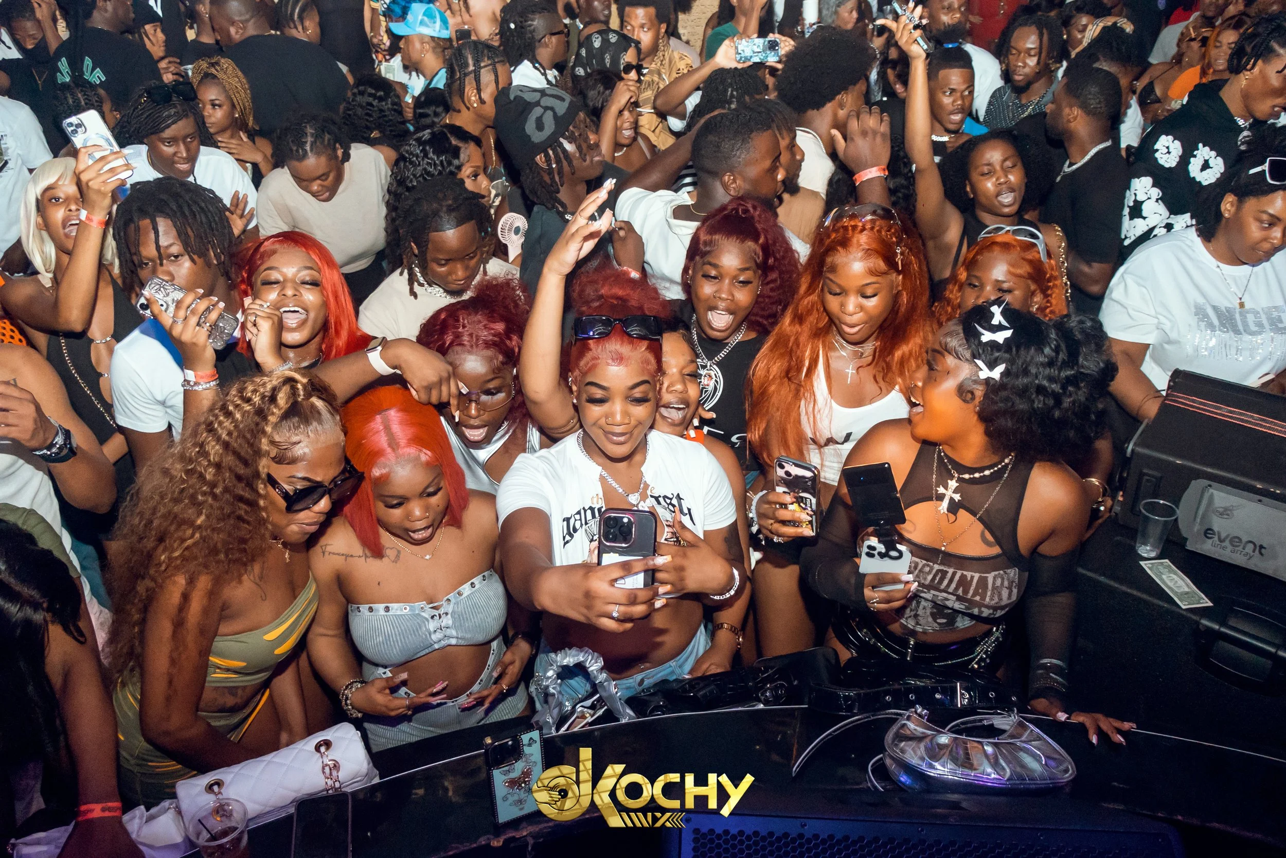 Crowd of people at a concert or party, many holding up phones to take pictures or videos. The crowd is lively and diverse, with women in the front smiling and dancing, some with brightly colored hair and accessories.