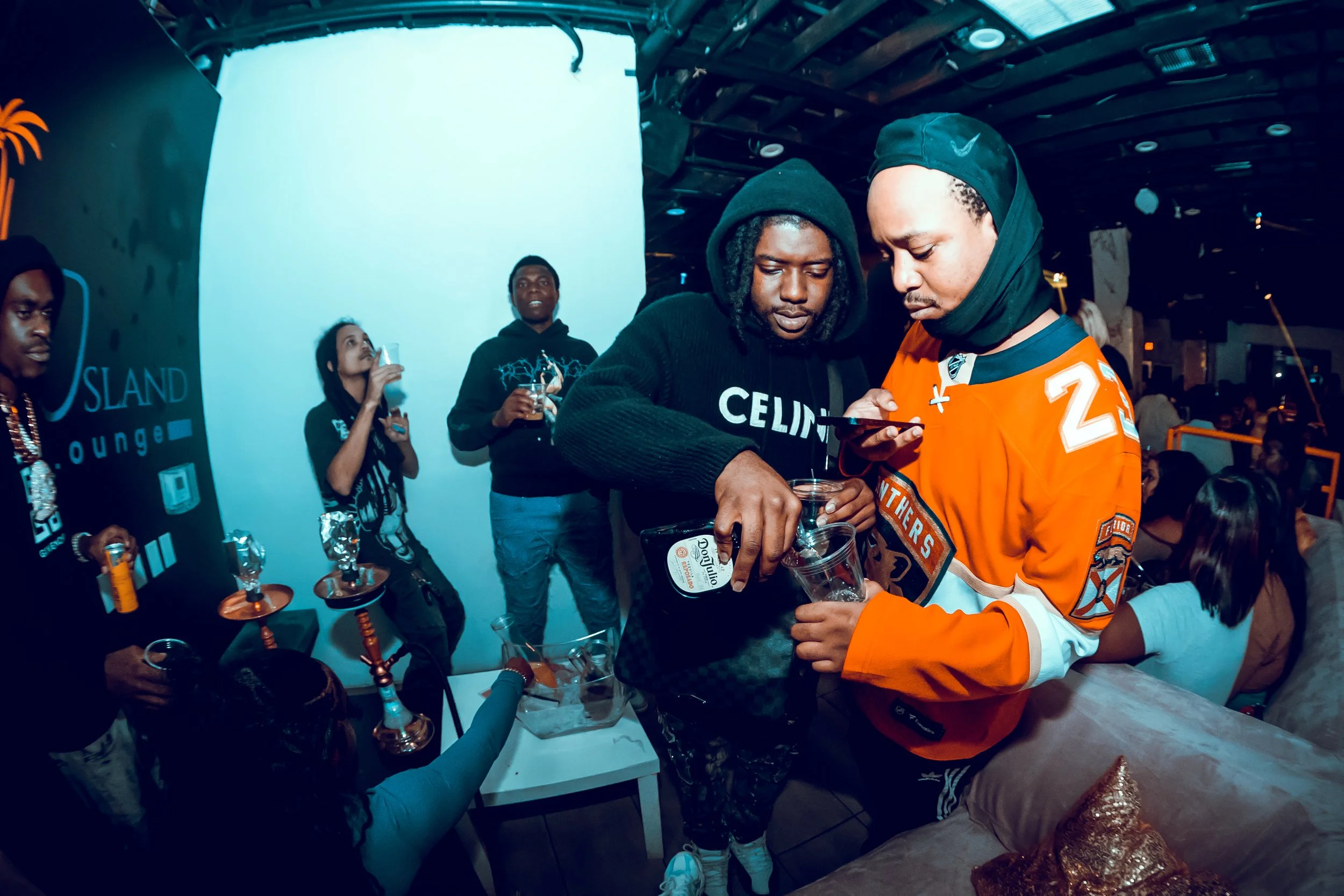 People at a party or lounge, some holding drinks, with two men in the foreground pouring drinks and looking at a phone, surrounded by others relaxing and socializing.