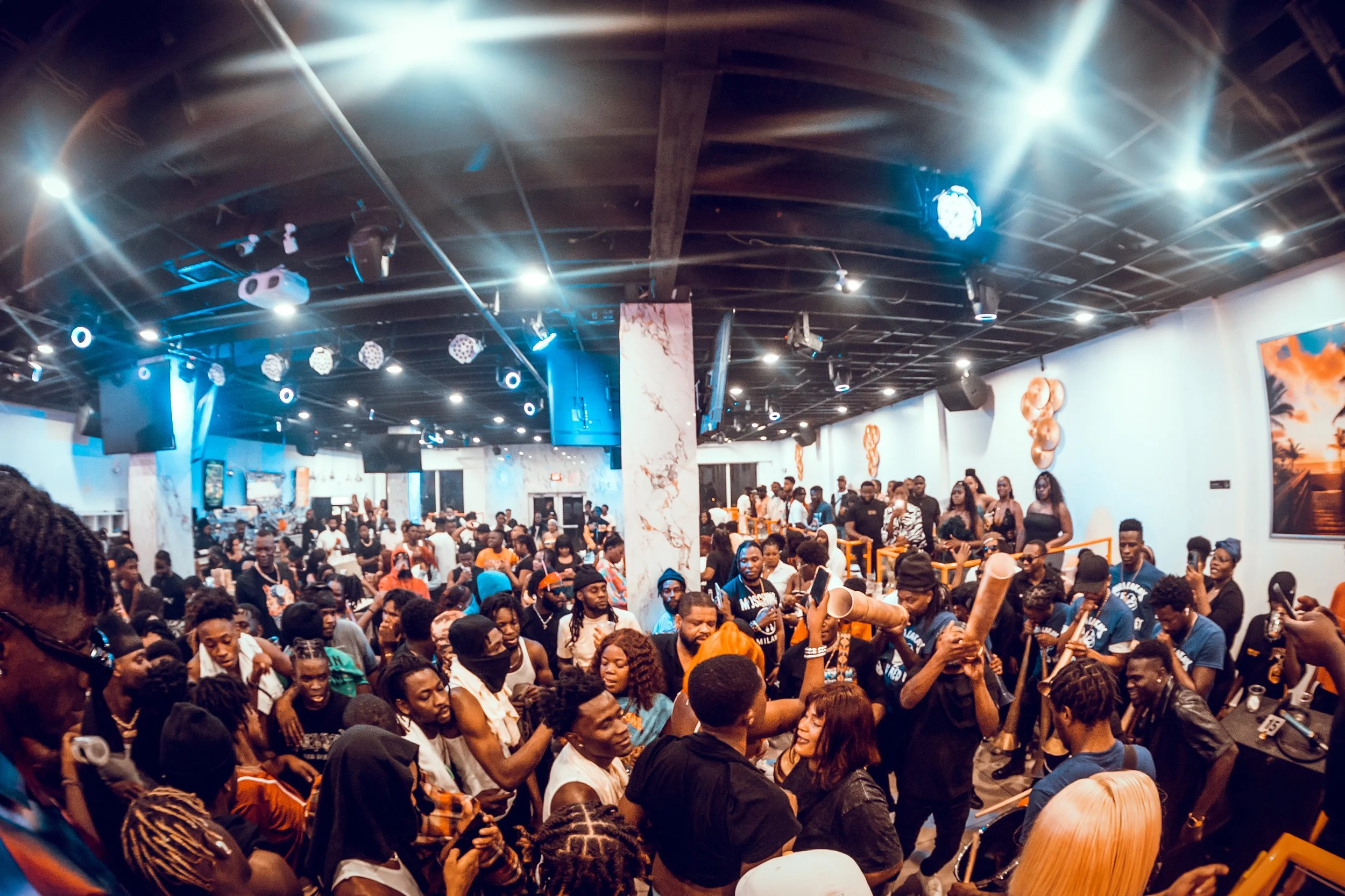 Crowd of people gathered in a lively indoor event space with modern decor, including black ceiling with lighting rigs and orange accents on the walls.