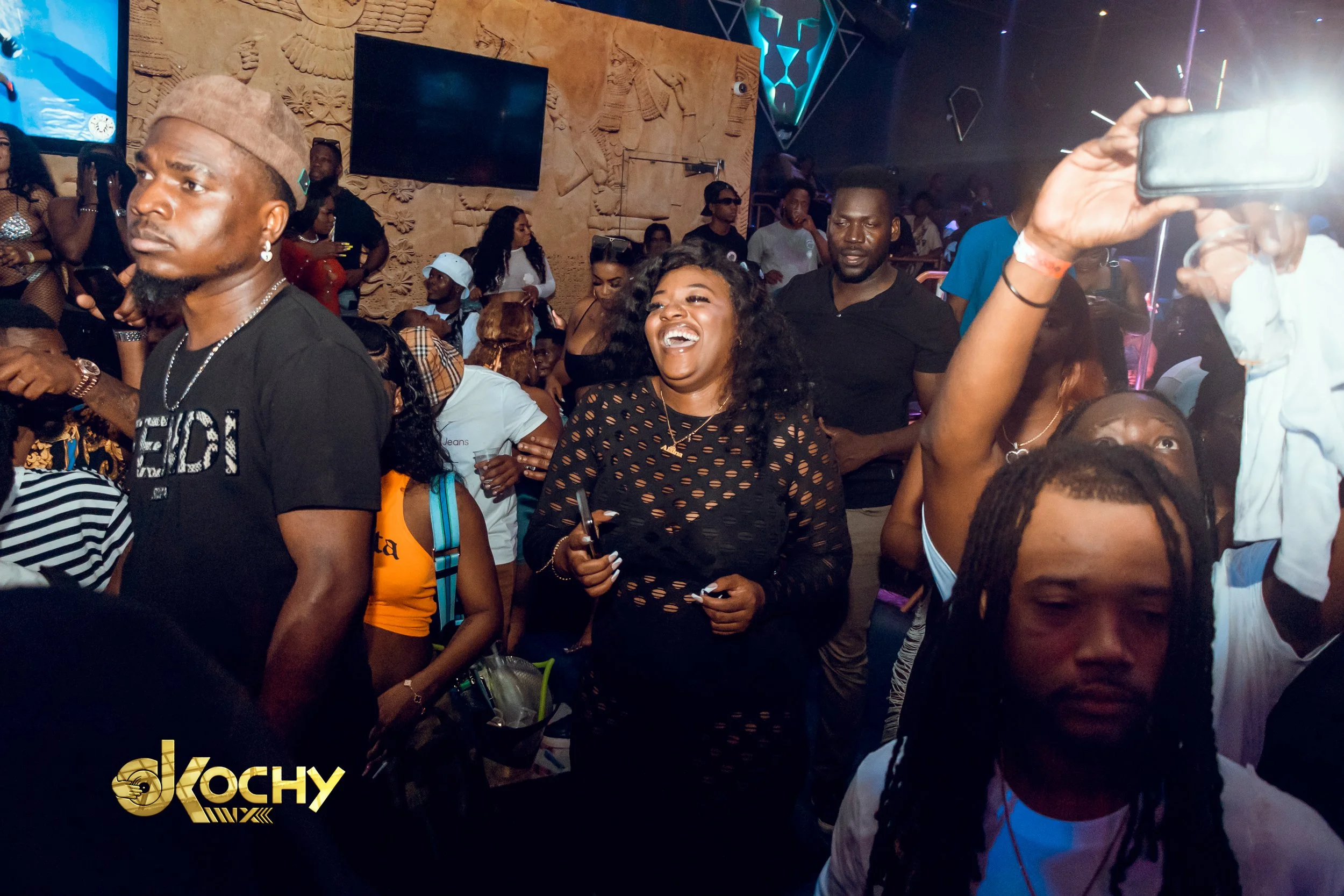 A lively nightclub scene with people dancing and enjoying themselves. A woman in the center is smiling and laughing, wearing a black, cut-out dress. Surrounding her are other club-goers, some with phones in hand, and a dark background with decorative