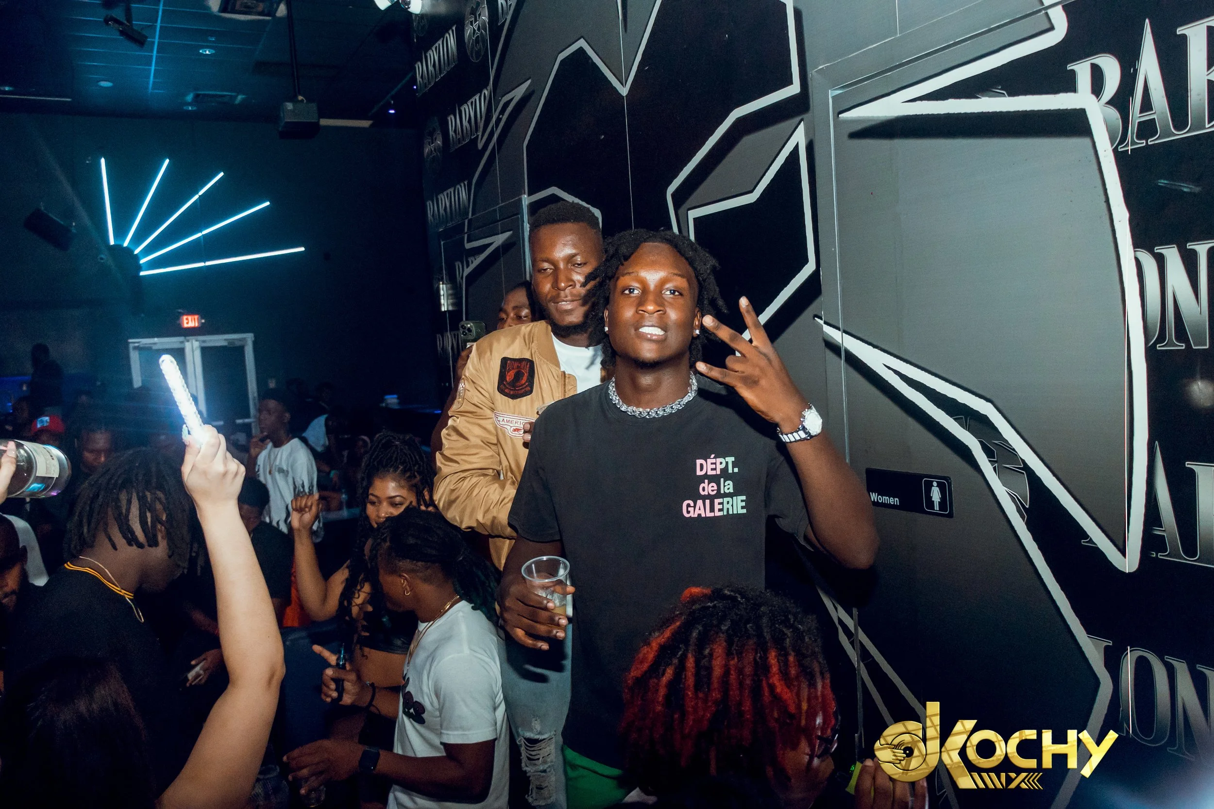 Group of young people at a nightclub or party, some dancing and taking pictures, with a dark background and neon lights.