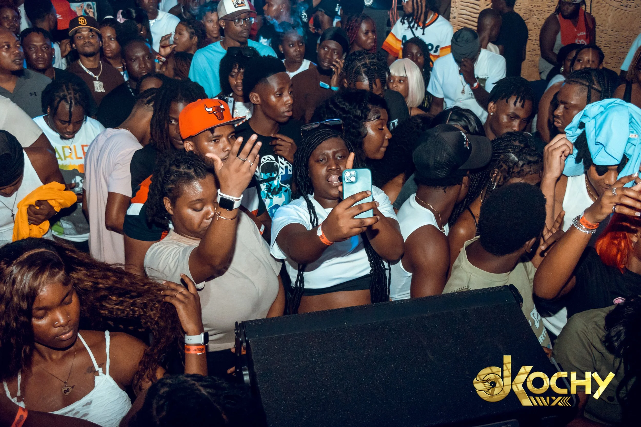 A crowd of people at a concert or event, many looking towards the stage, some taking photos or videos, with the DJ Kochy logo in the bottom right corner.