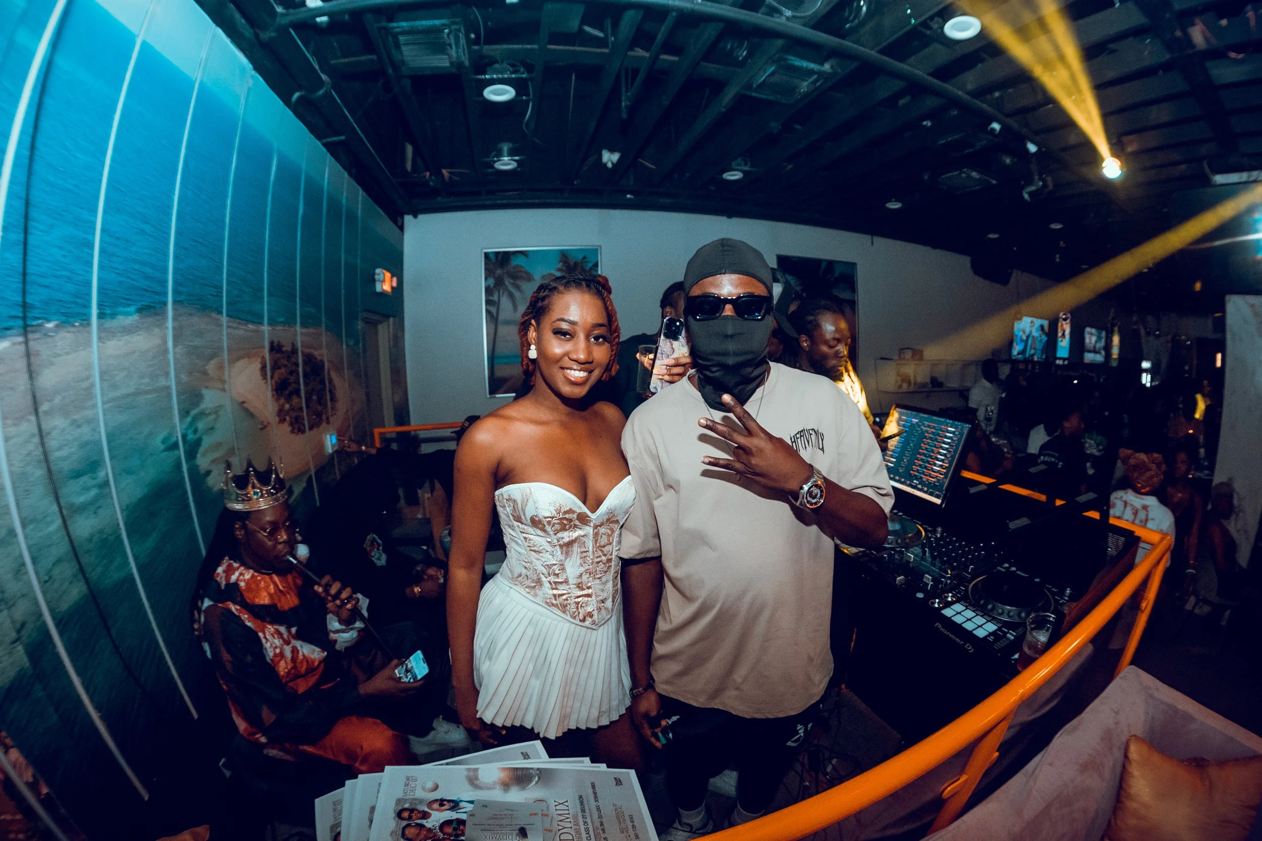 A woman in a white dress smiles next to a DJ wearing sunglasses, a mask, and a cap at a party or event with a DJ setup and other people in the background.