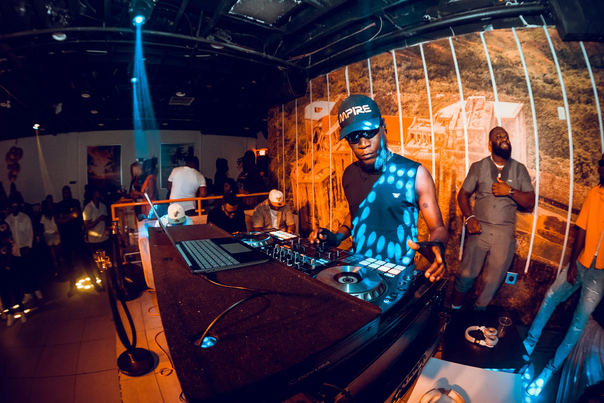 A DJ wearing sunglasses, a hat, and a sleeveless shirt, is spinning music at a party with a crowd dancing and enjoying the event. The background features a large mural of a scenic landscape.
