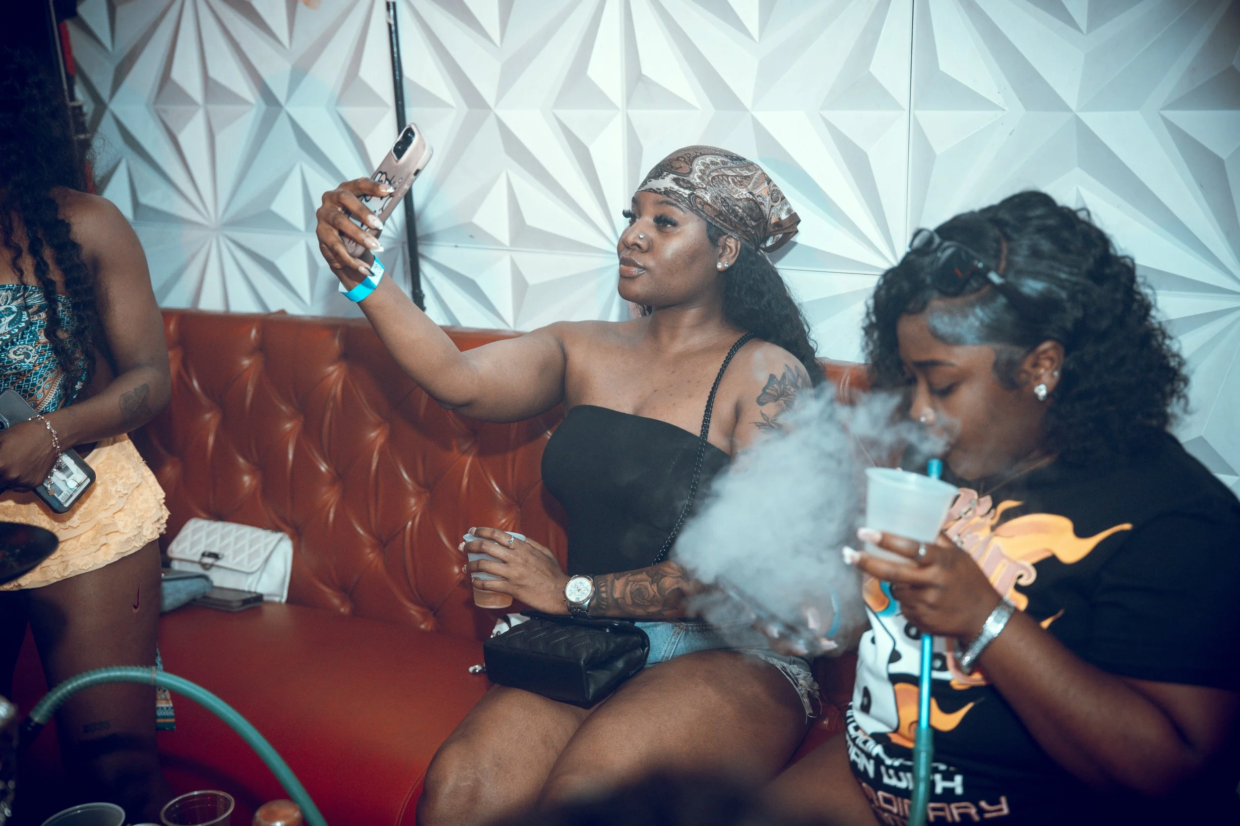 Two women sitting on a red couch at a social gathering, one taking a selfie with a smartphone, the other smoking hookah, with a modern geometric-patterned wall in the background.