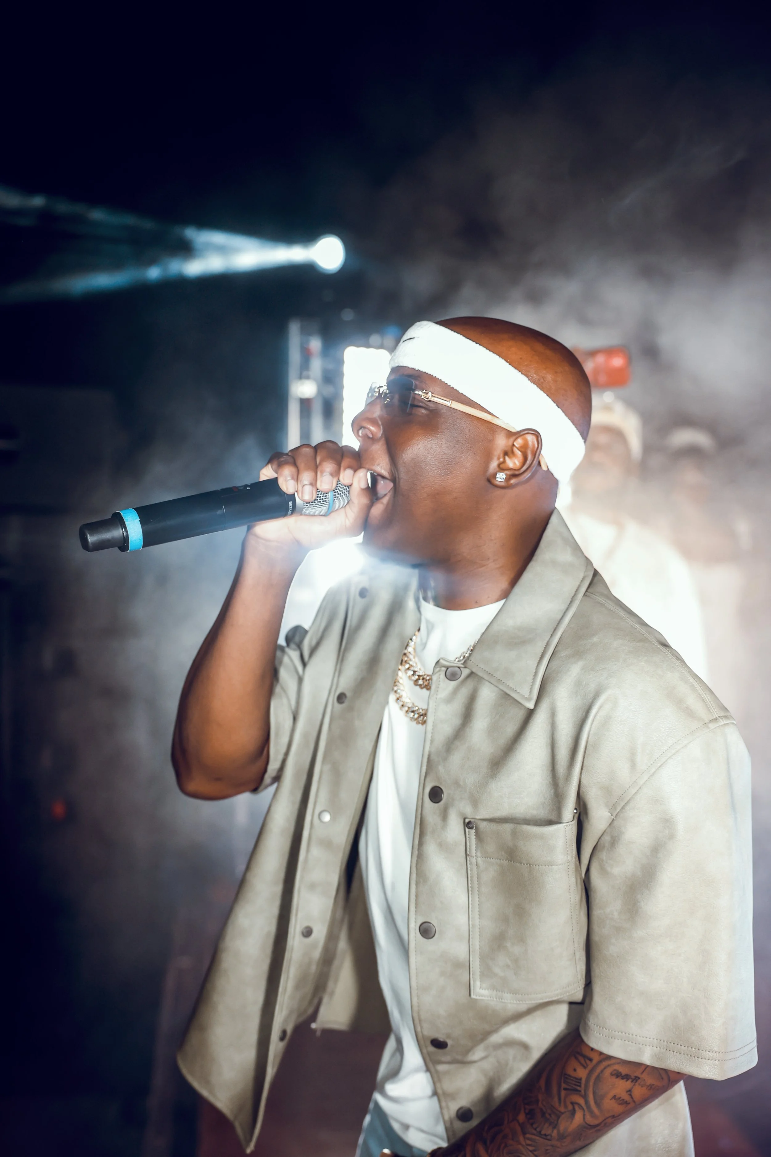A male rapper performing on stage with a microphone, wearing a white headband, sunglasses, a beige jacket, and jewelry.