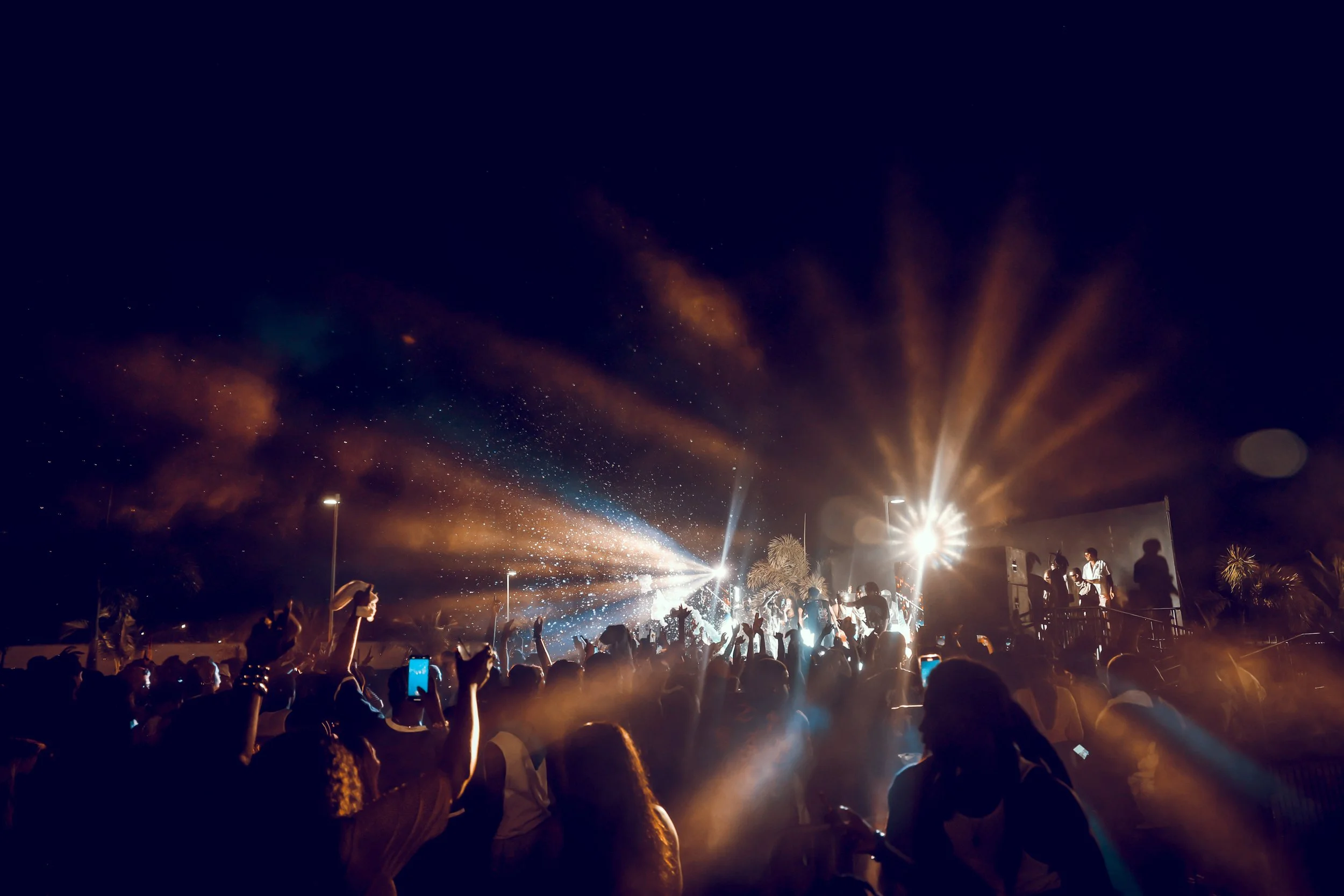 Large crowd at an outdoor concert or music festival at night, with stage lights, screens, and smoke effects, people dancing and taking photos.