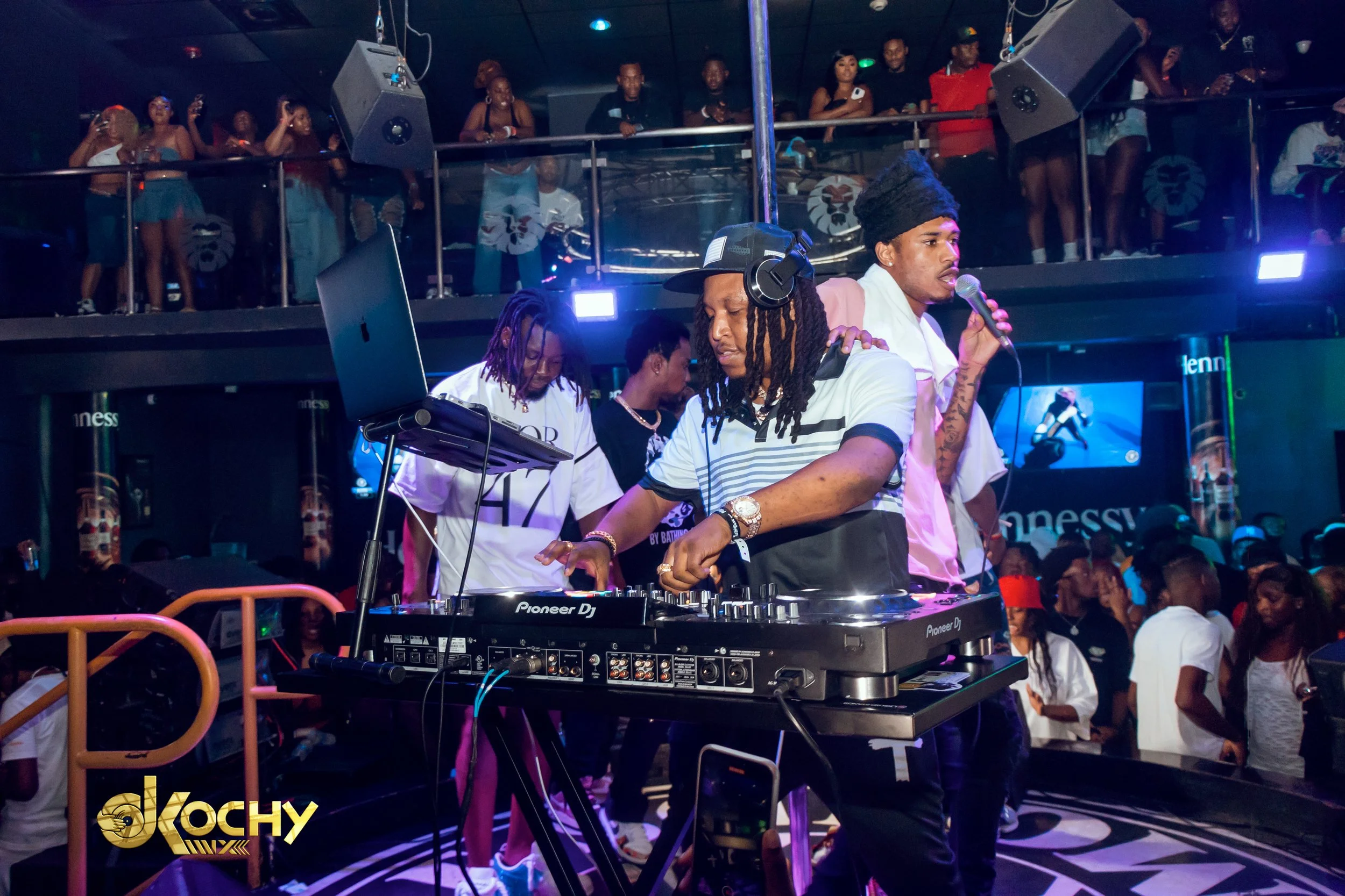 A DJ wearing a cap and wristwatch is mixing music on a Pioneer DJ controller at a nightclub. Beside him, a man with an afro and a white shirt is holding a microphone. The background features a crowd of people dancing and watching the performance from
