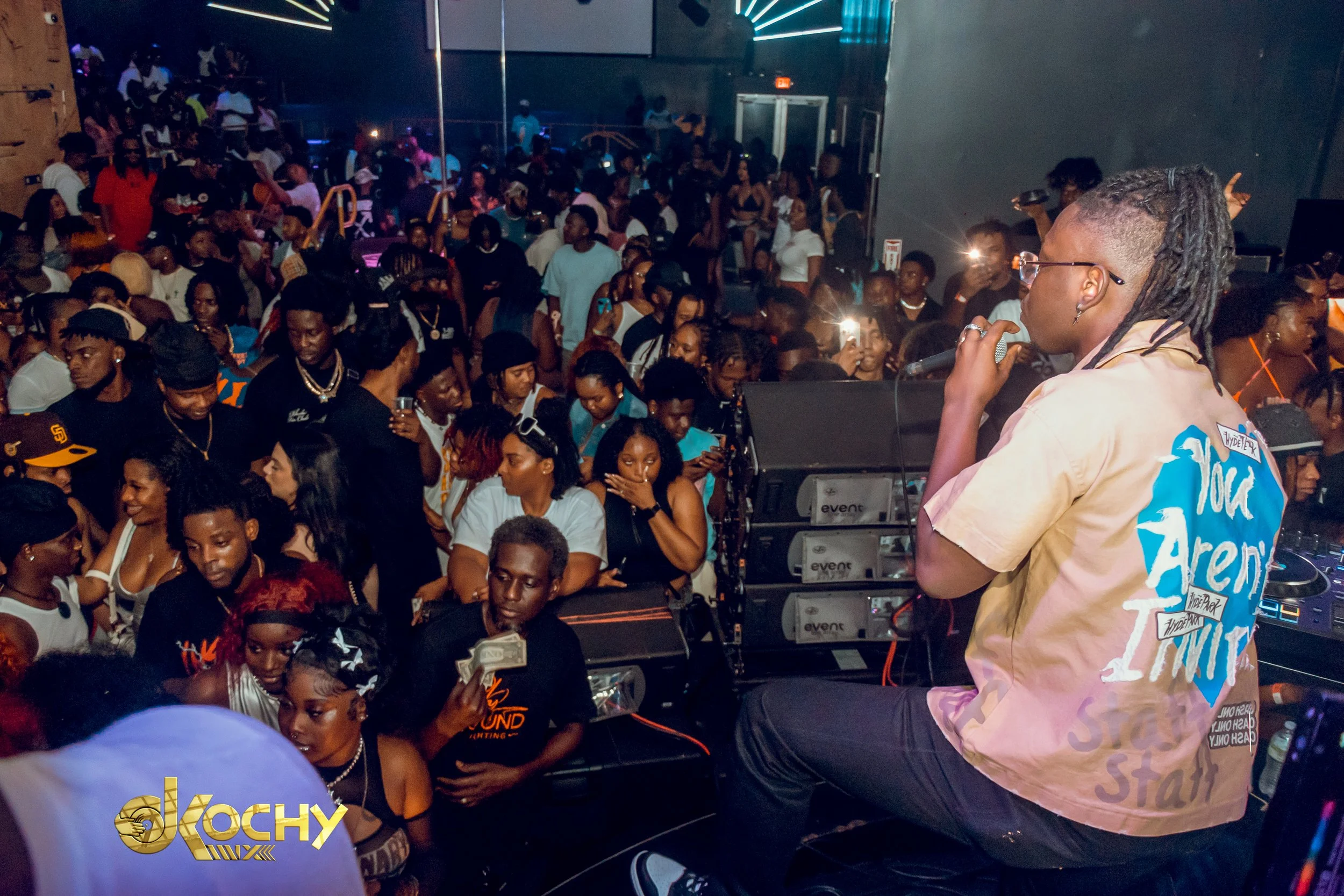 A DJ with dreadlocks and glasses performing on stage at a lively house music event, with a large crowd dancing and taking photos in a club-like setting.