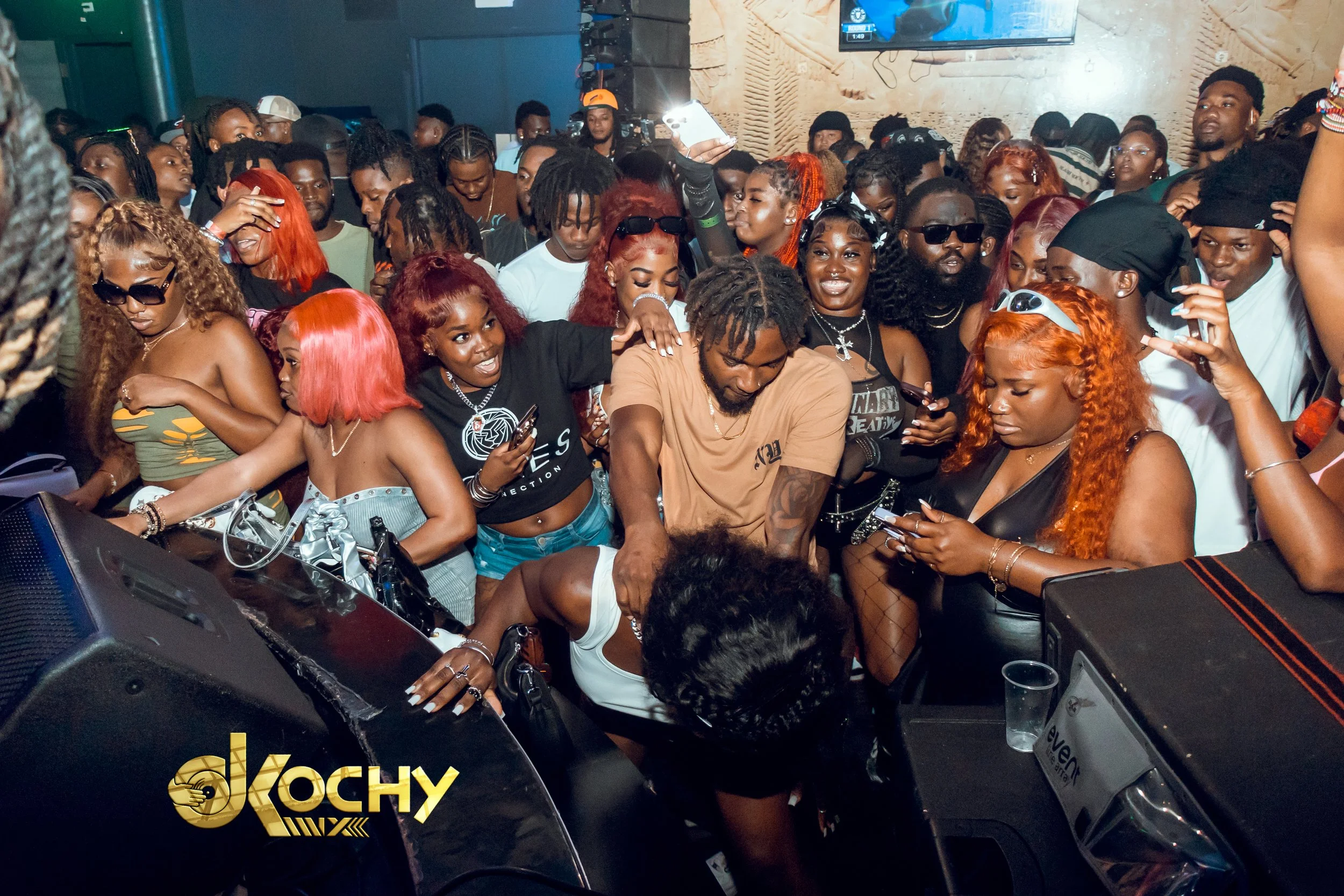 A crowded indoor music event with a diverse group of people dancing and enjoying the performance. The scene includes a DJ, colorful hairstyles, and many people holding phones capturing the moment.