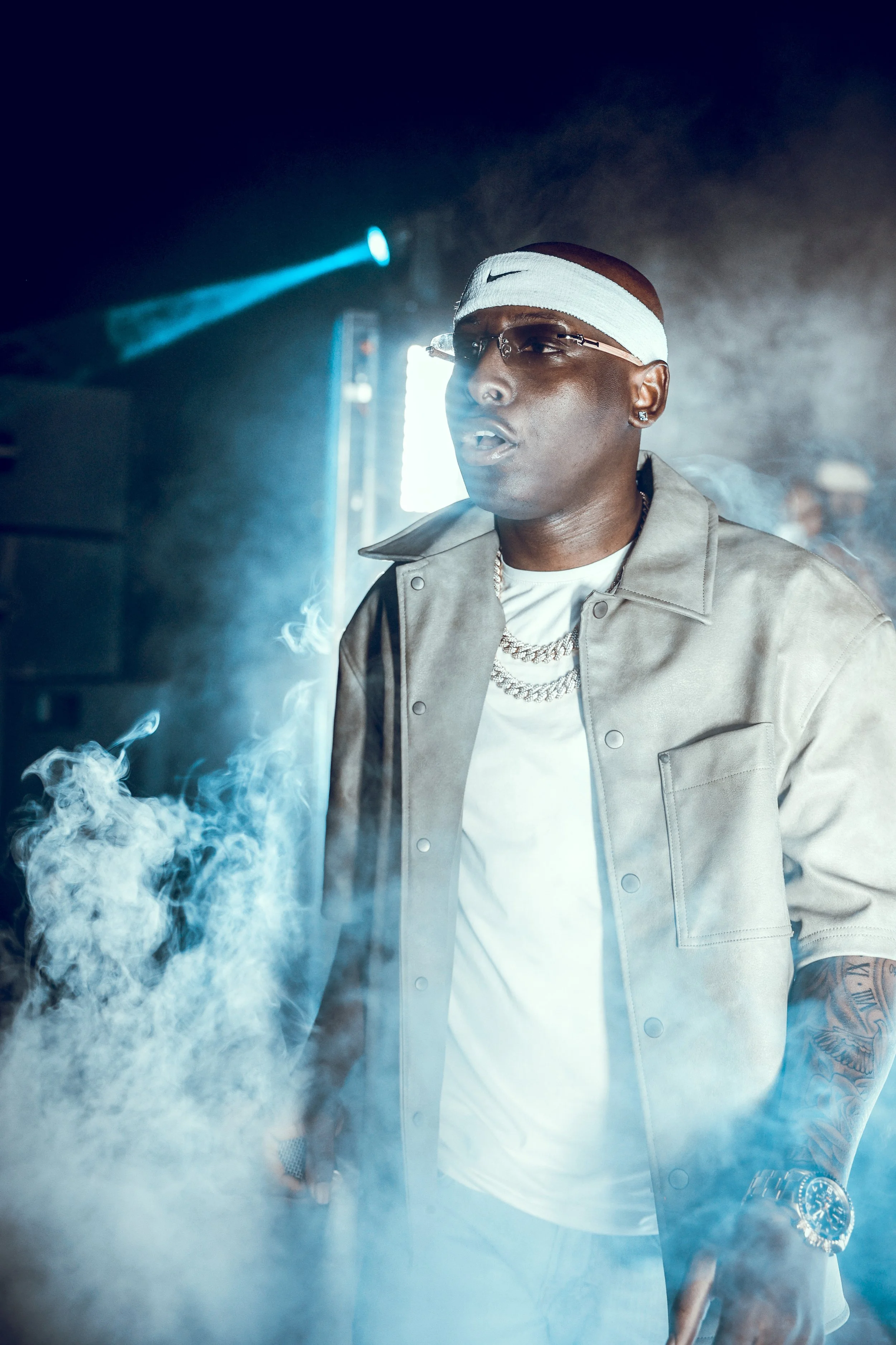 A man dressed in a beige jacket, white t-shirt, white headband, sunglasses, and jewelry standing in a smoky setting with blue lighting.