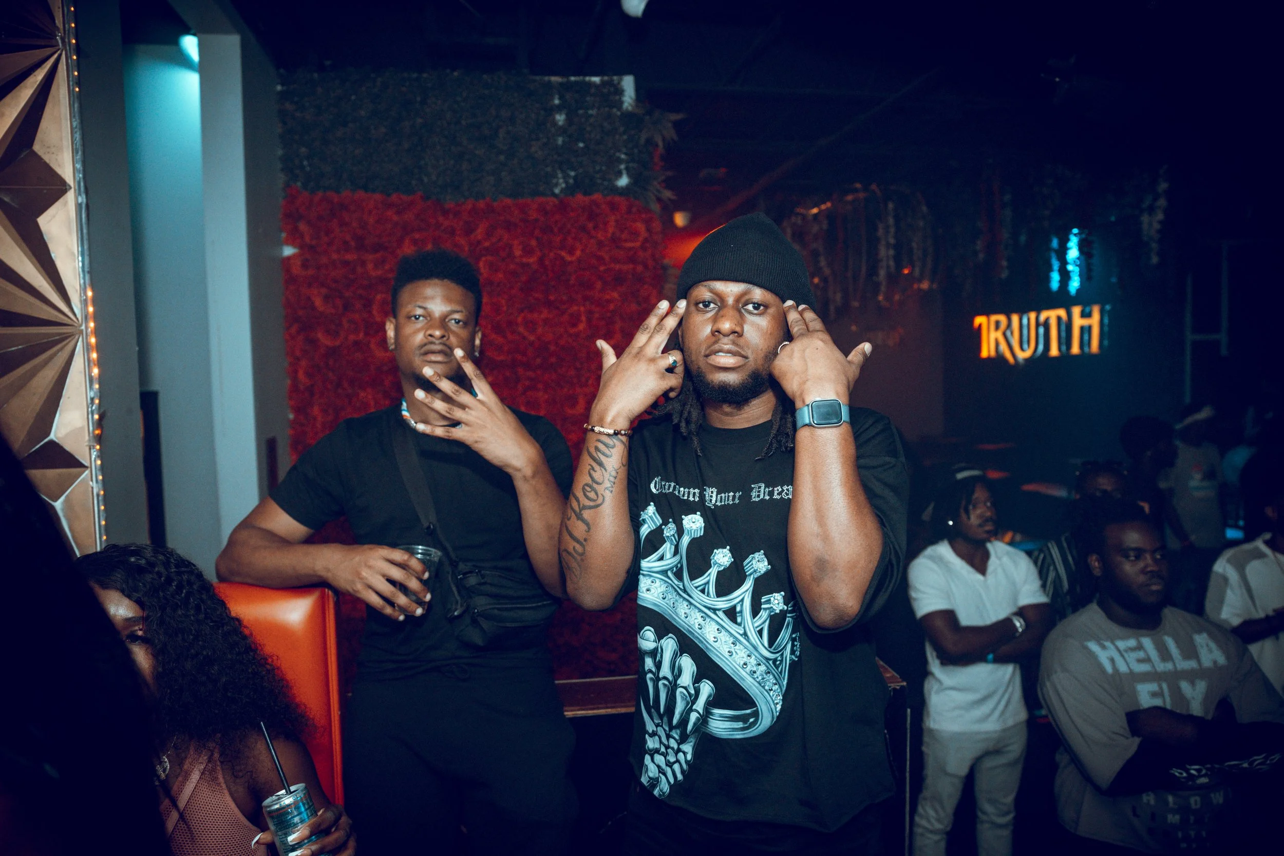 Two men posing at a nightclub with a red wall and dark background. One man is wearing a black beanie, a tattooed arm, and making a gesture near his ears. The other man is holding a drink, has short hair, and a camera strap around his neck. There are 