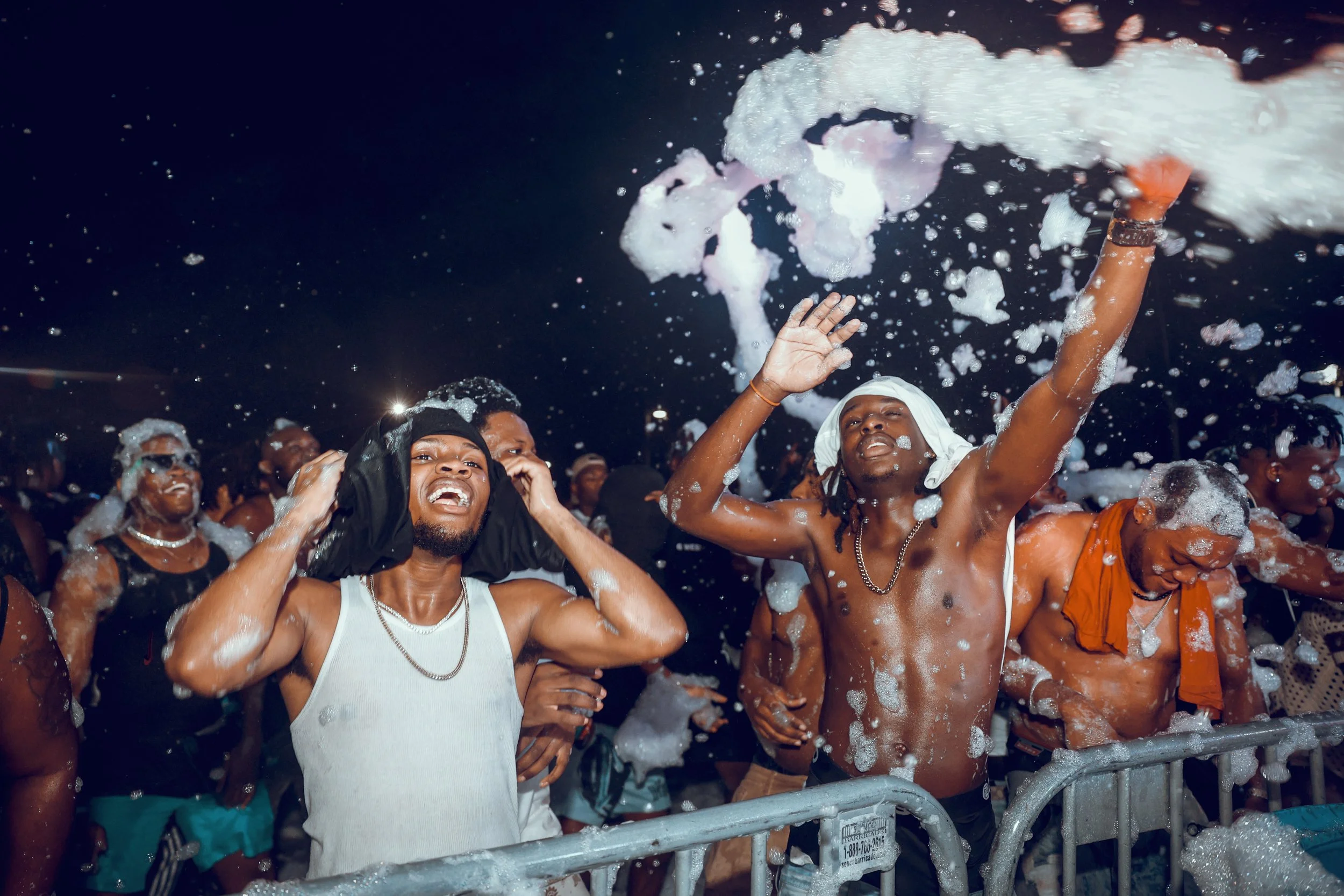 People dancing and smiling at a foam party, with foam falling from above and crowd in the background.