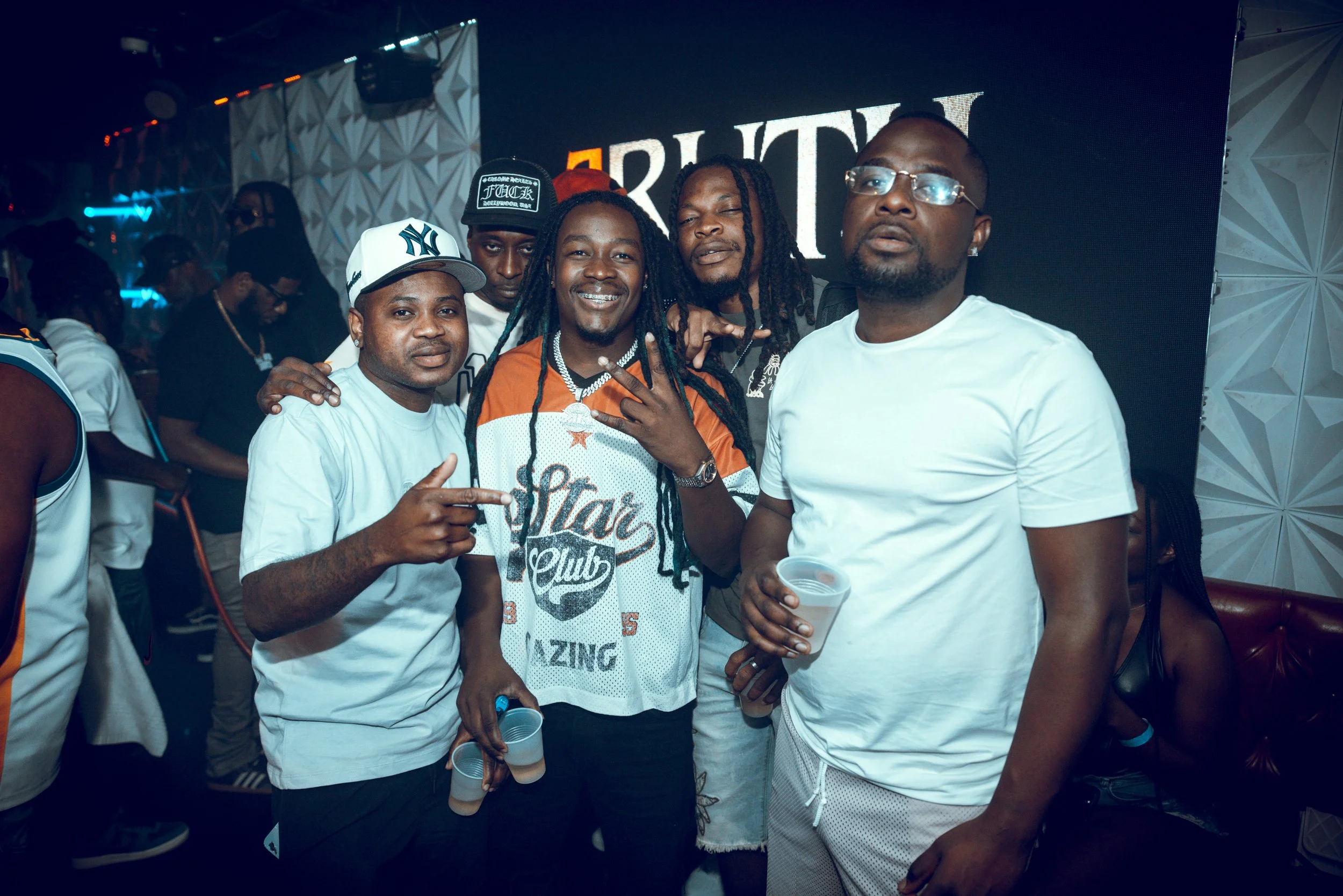 Group of men at a party or event, some holding drinks, with a large screen displaying the word 'BRUTAL' in the background.
