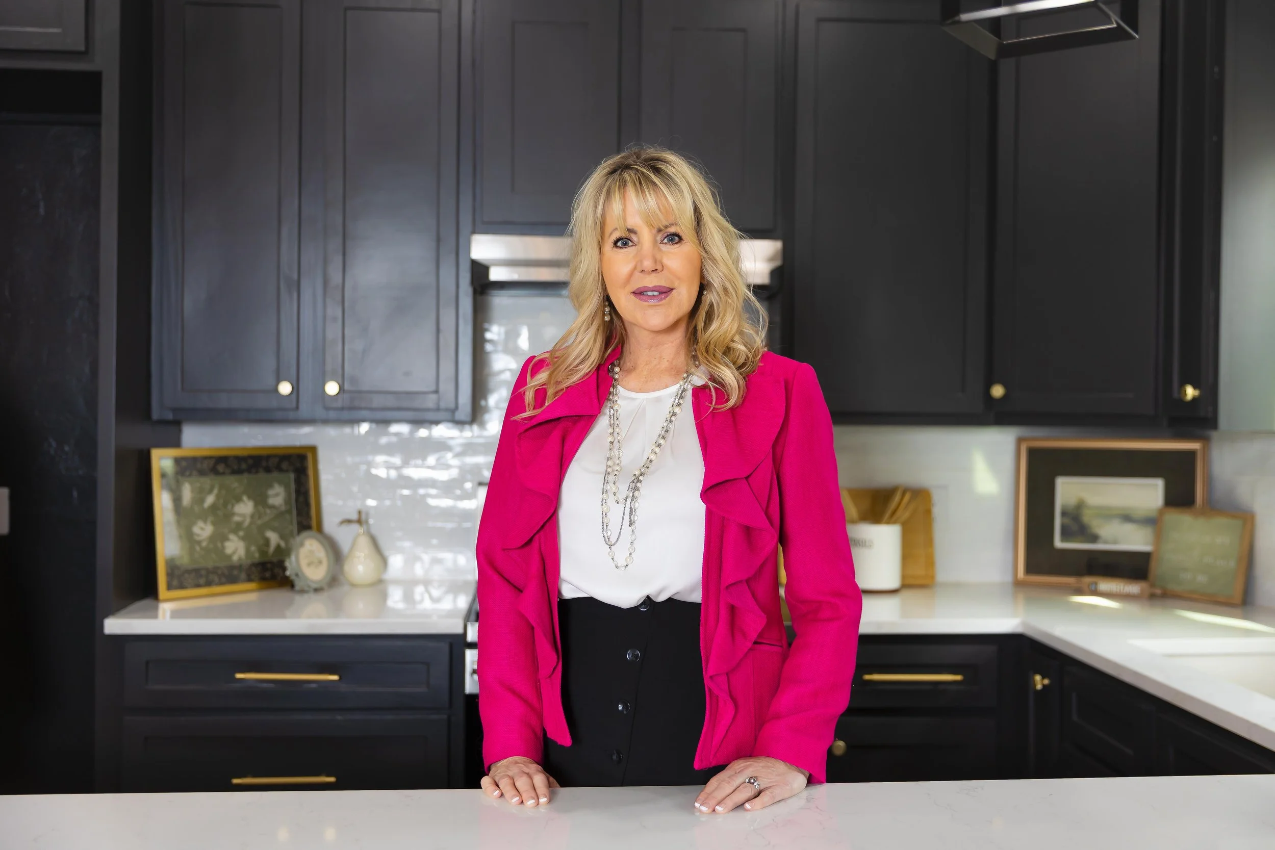 A woman with blonde hair wearing a hot pink blazer and jewelry standing in a modern kitchen with black cabinets and a white countertop.
